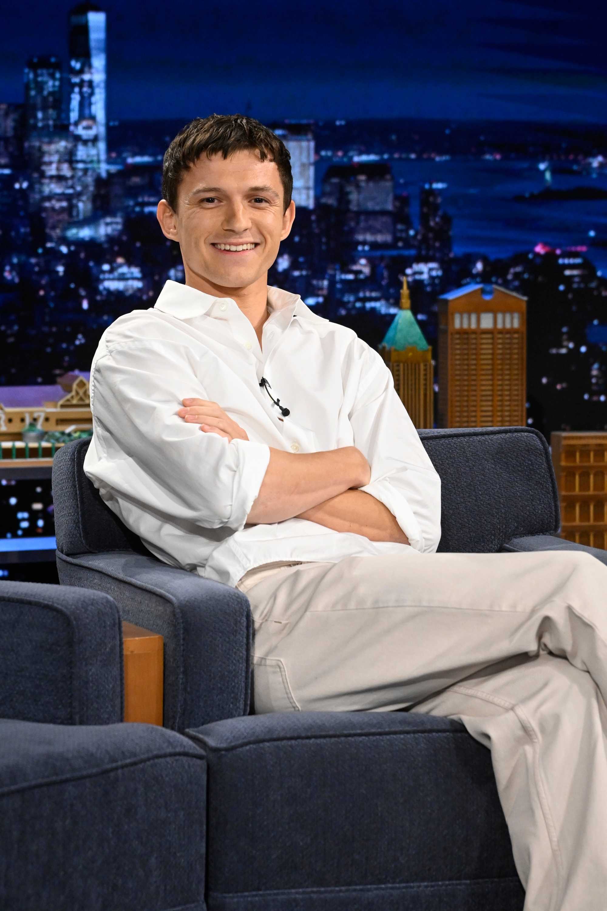 Person in white shirt and light pants smiling, sitting on a talk show set with a cityscape in the background