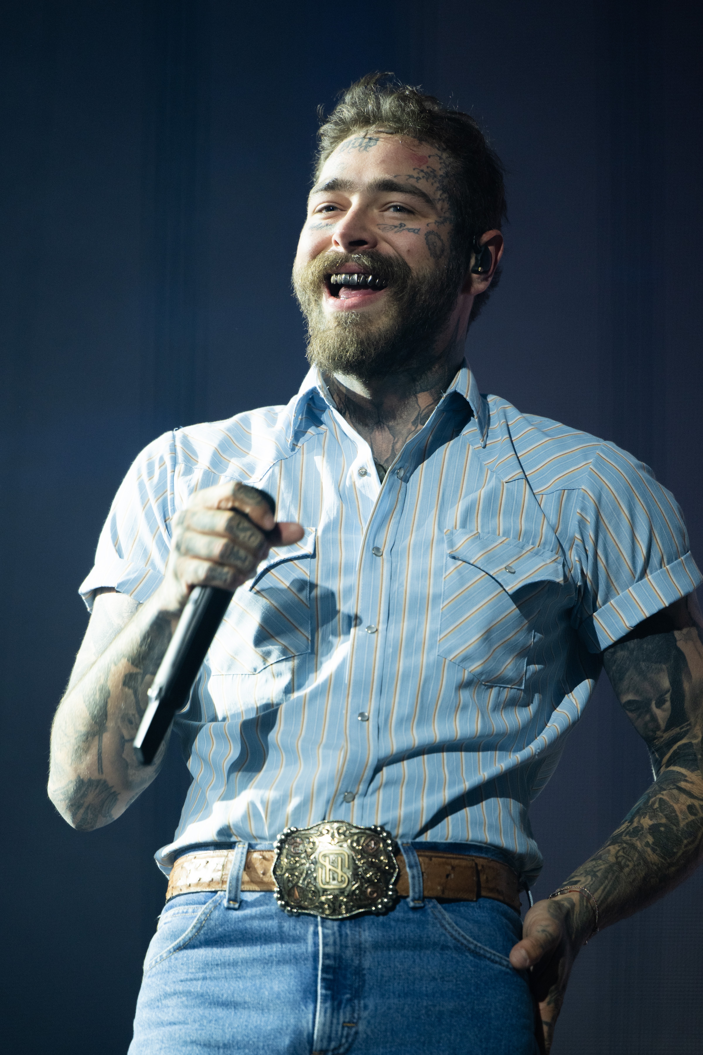 A performer with tattoos sings passionately on stage, wearing a striped shirt, jeans, and a large belt buckle