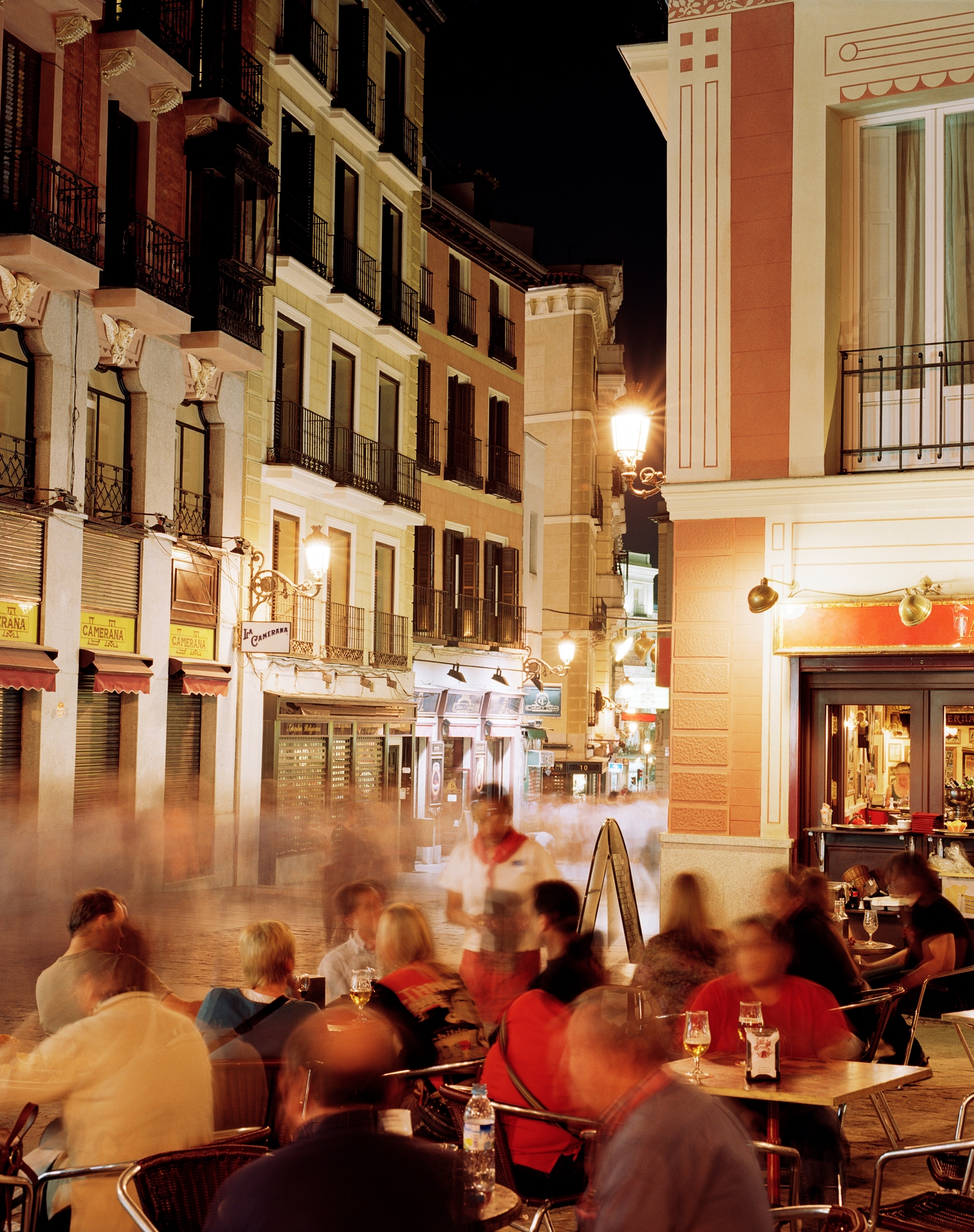 People dining at outdoor tables on a bustling city street at night, with blurred motion suggesting conversation and activity