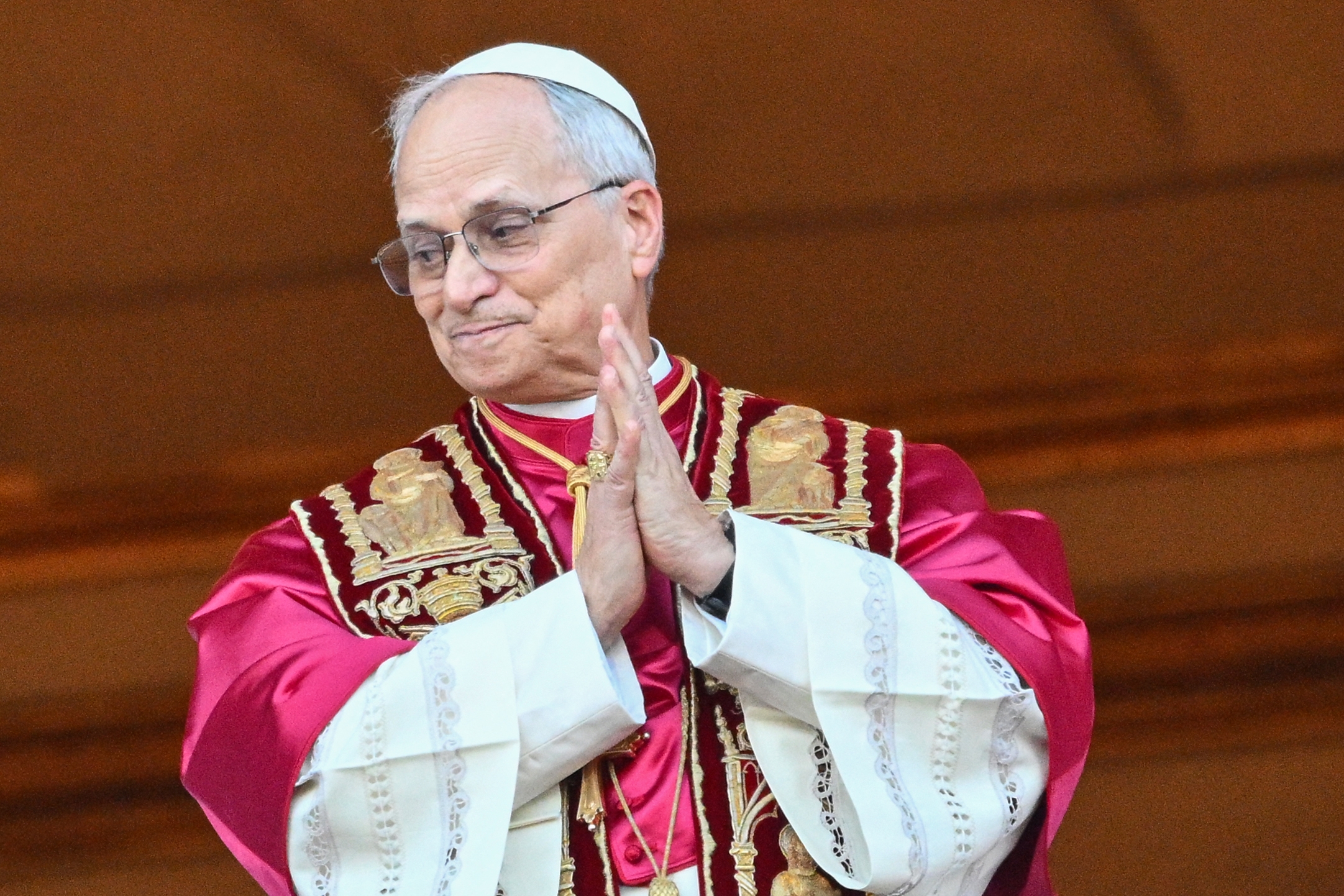 A man dressed in ceremonial robes and a white skullcap stands with folded hands in a formal setting