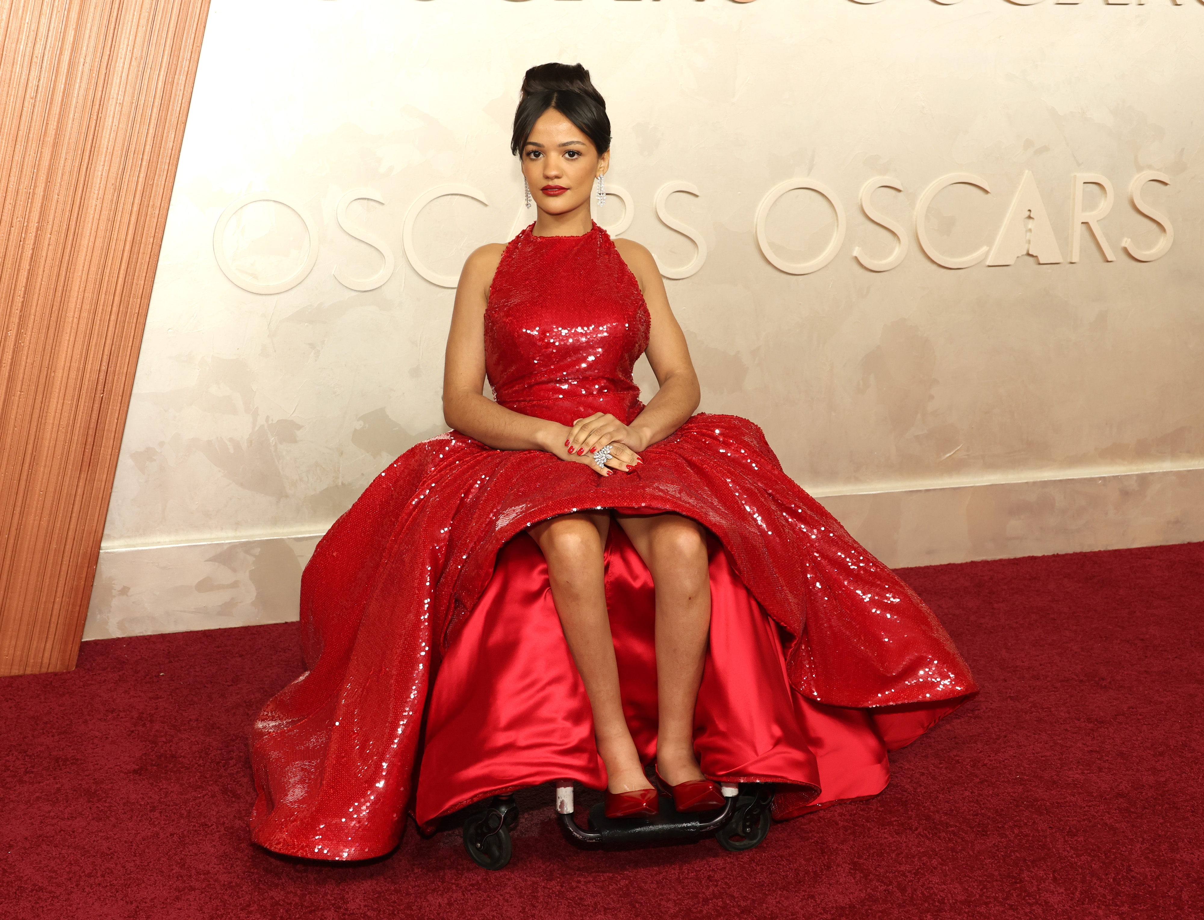 Person in a sequined gown with a high neckline at a red carpet event, seated on a mobility device. "OSCARS" is visible on the backdrop