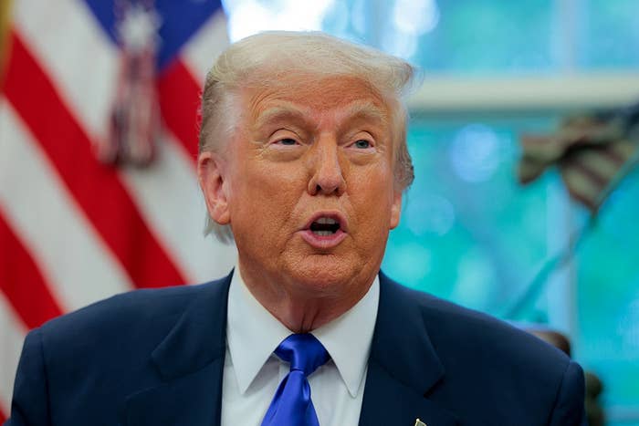 A person in a suit and tie in front of a blurred American flag background, speaking with a focused expression