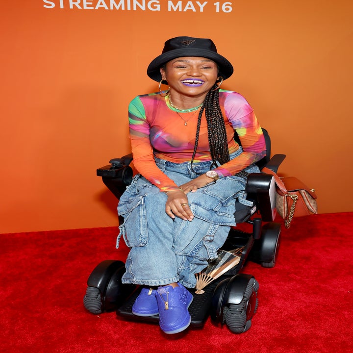 Person in wheelchair on red carpet wearing a vibrant top, denim cargo pants, and a hat. They are smiling joyfully