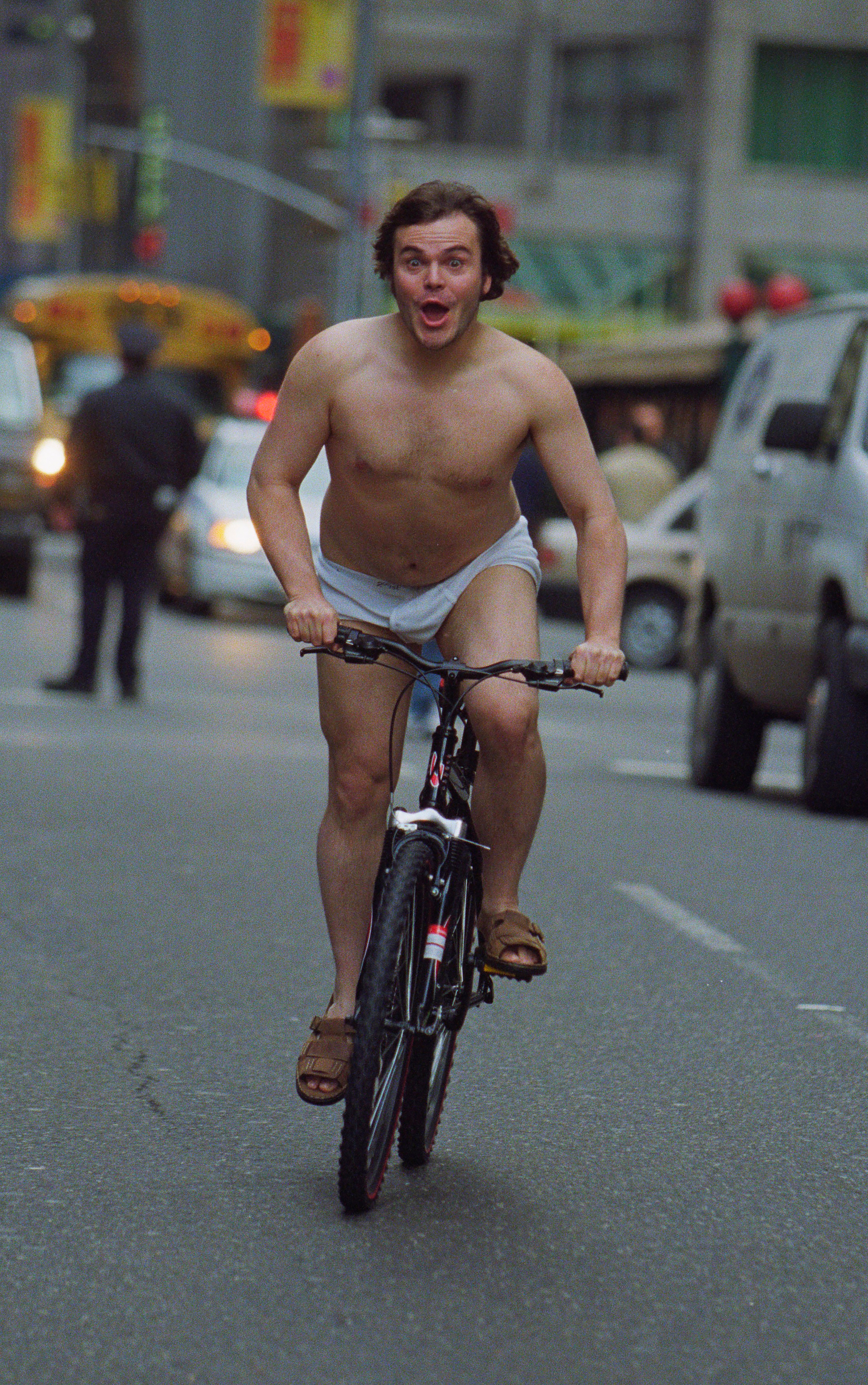 A person rides a bicycle on a city street wearing only shorts and sandals, with a surprised expression