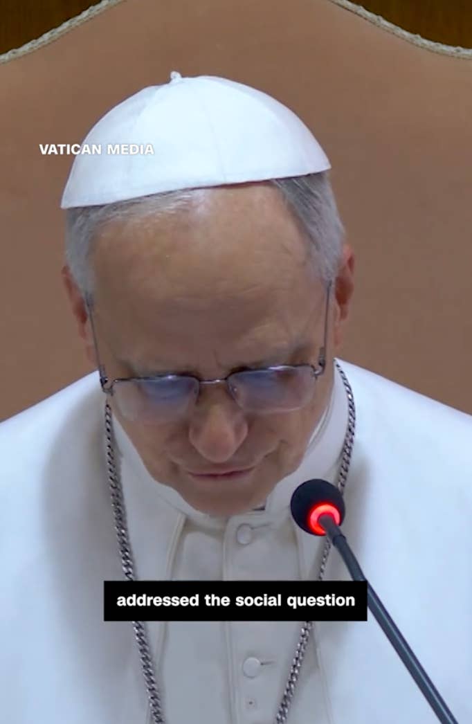 A religious leader in white attire, speaking into a microphone during an event, with a caption about addressing social issues