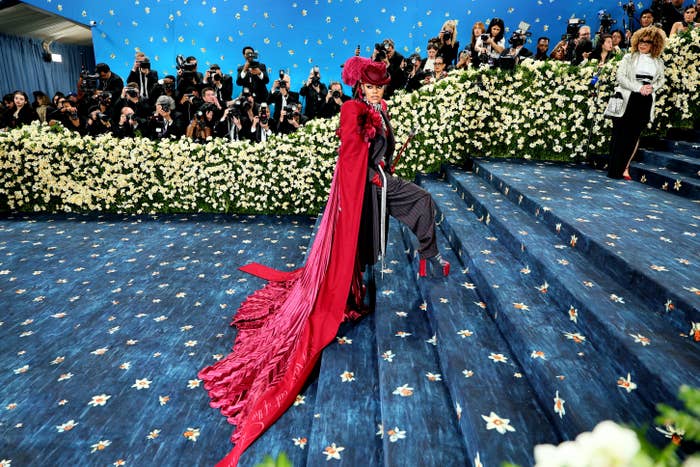 Celebrity on a grand staircase, wearing an extravagant red gown with a long train, surrounded by photographers and floral decorations