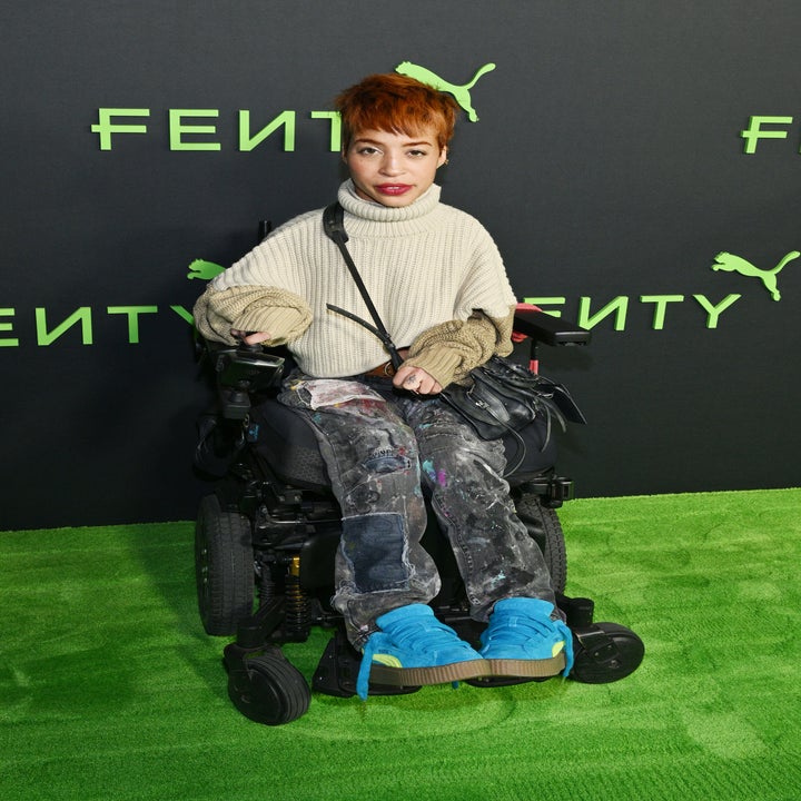 Person seated in a wheelchair, wearing a turtleneck sweater and splatter-paint pants, at a Fenty event