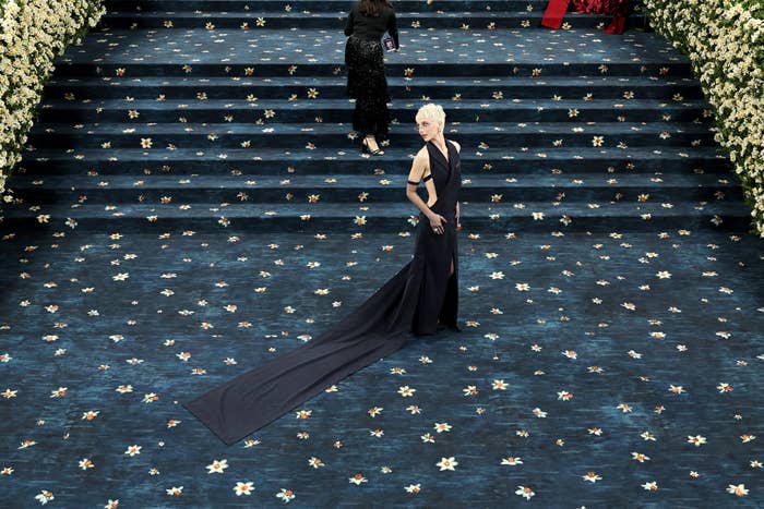 Person in an elegant, flowing gown poses on steps adorned with floral designs, surrounded by white flowers, at a formal event