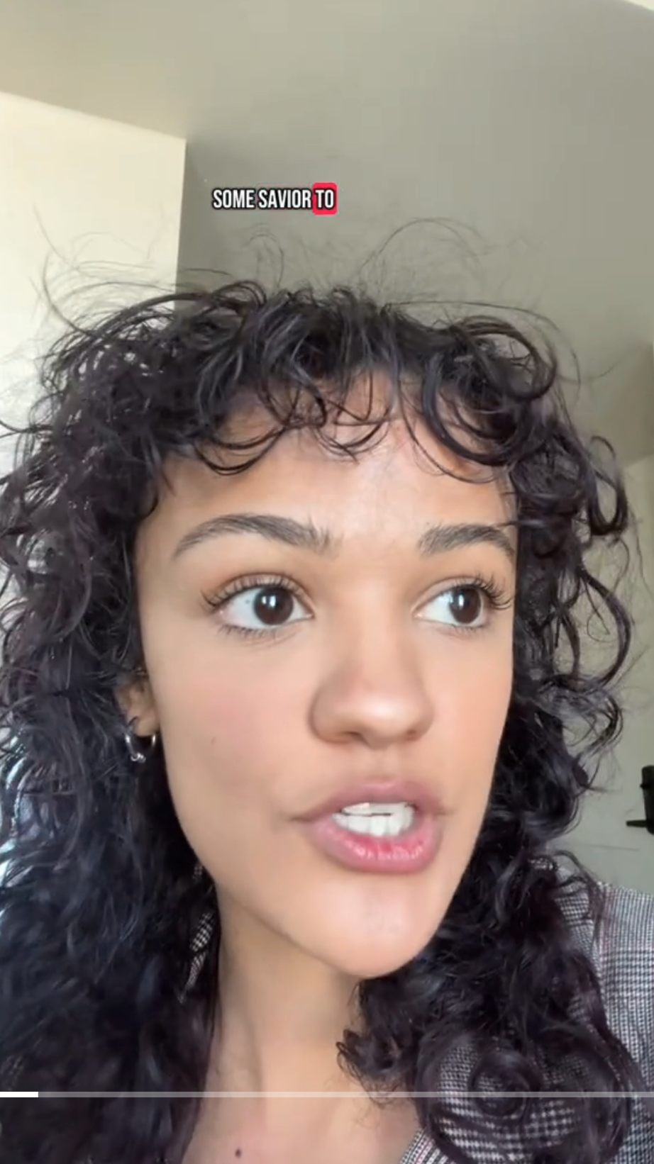 Person speaking with expressive gaze, short curly hair, and wearing earrings. Background is indoors. Text above reads: "Some savior to"
