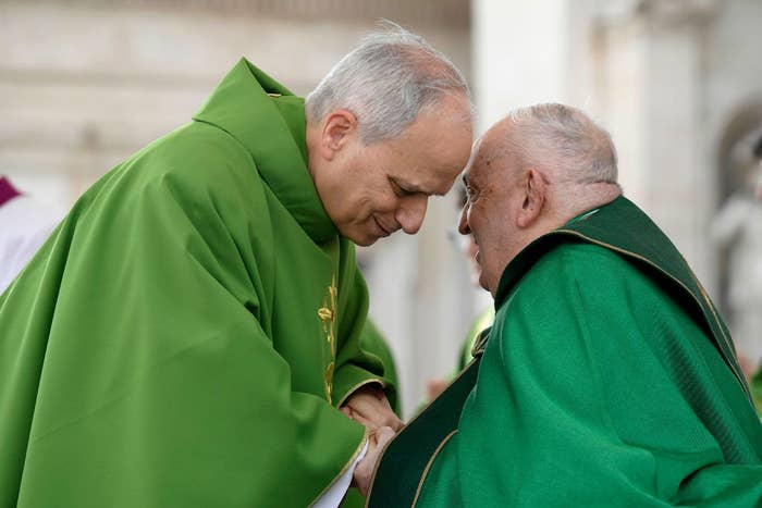 Two elderly men, one bowing, wearing matching ceremonial robes, engaged in a respectful greeting or conversation at a formal event