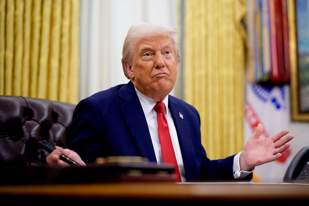 A person in a suit is sitting at a desk in an office, gesturing with both hands, with a neutral expression