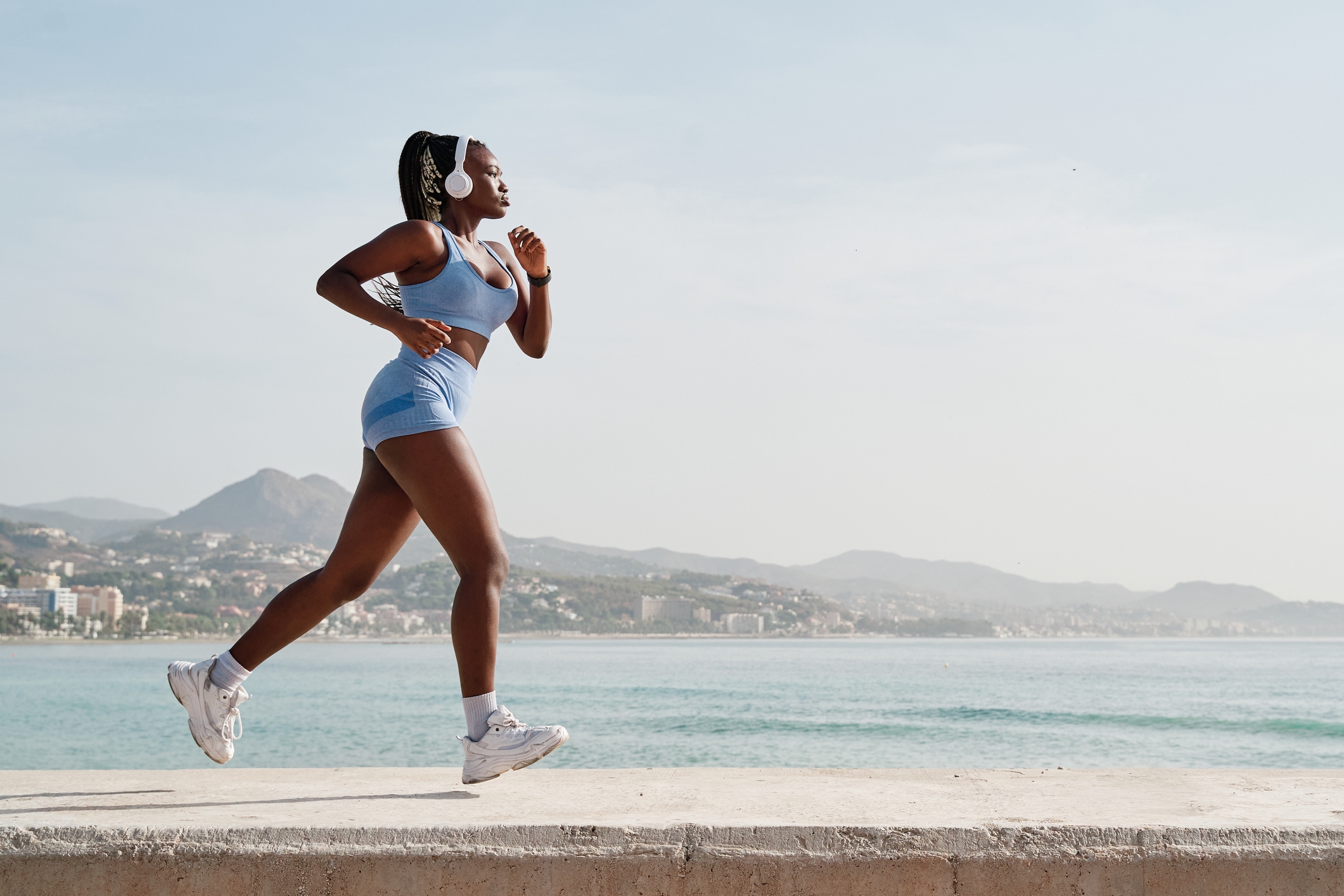 Person jogging by the sea in athletic wear, with mountains and cityscape in the background