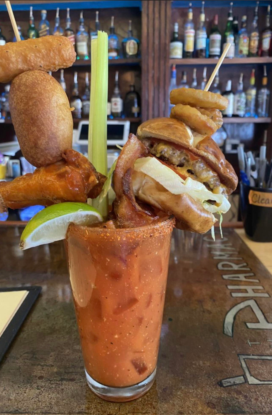 A Bloody Mary garnished with a slider, chicken wing, mini corn dog, onion rings, celery, bacon, and lime, set on a bar counter