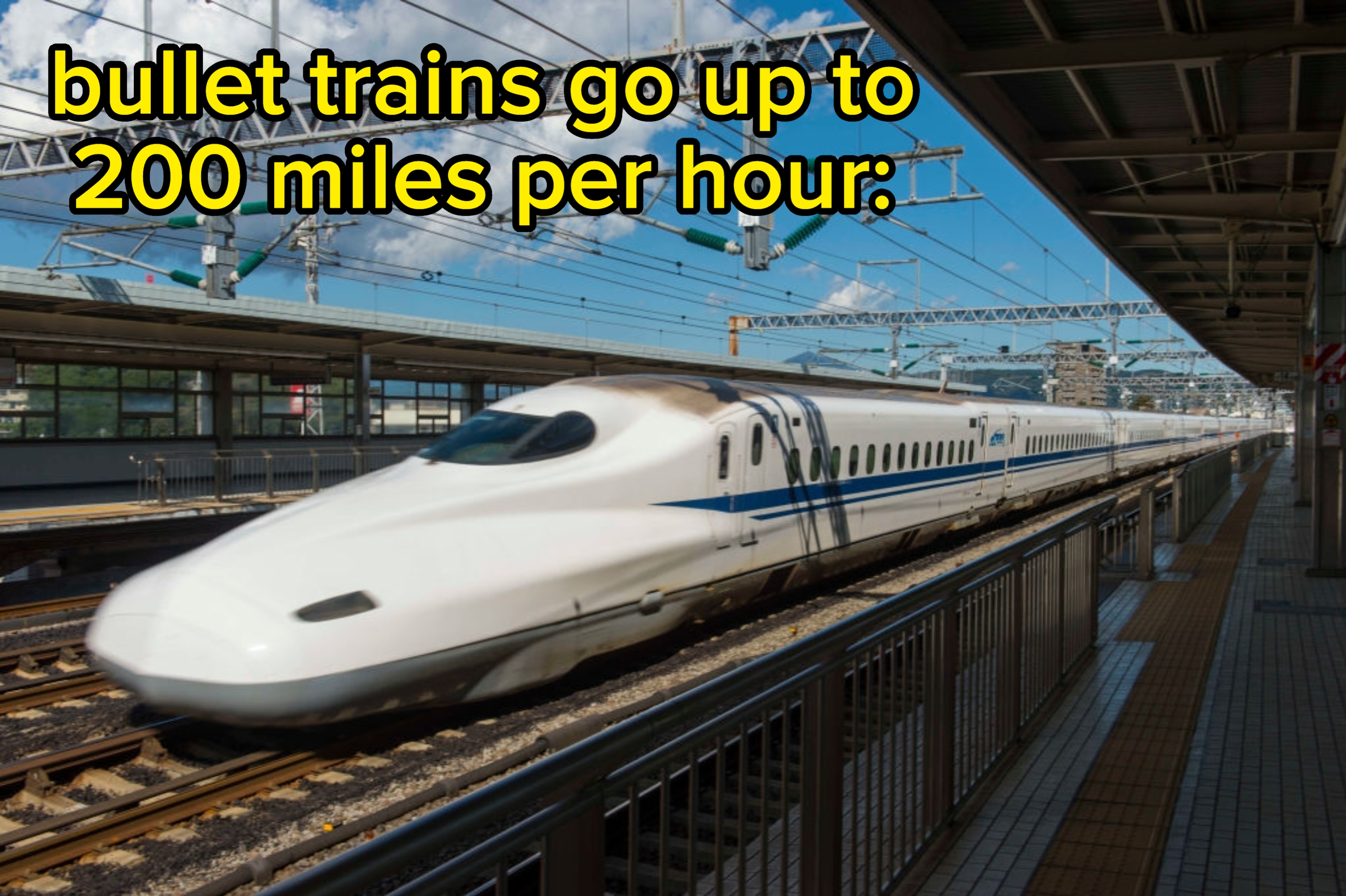 A high-speed bullet train travels quickly along railway tracks at a station, with overhead cables and a partly cloudy sky in the background