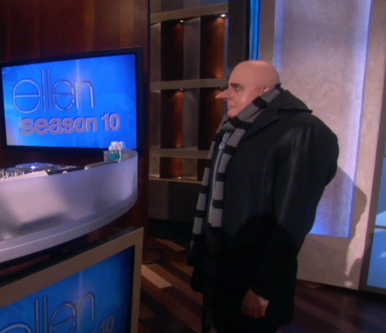 A person dressed as a character with a bald head and long nose, wearing a striped scarf and black coat, stands by a talk show set marked "Ellen Season 10."