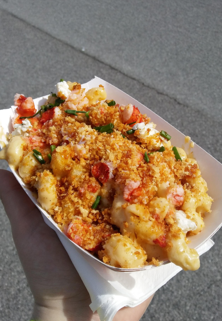 A hand holds a tray of lobster mac and cheese topped with breadcrumbs and herbs, outdoors