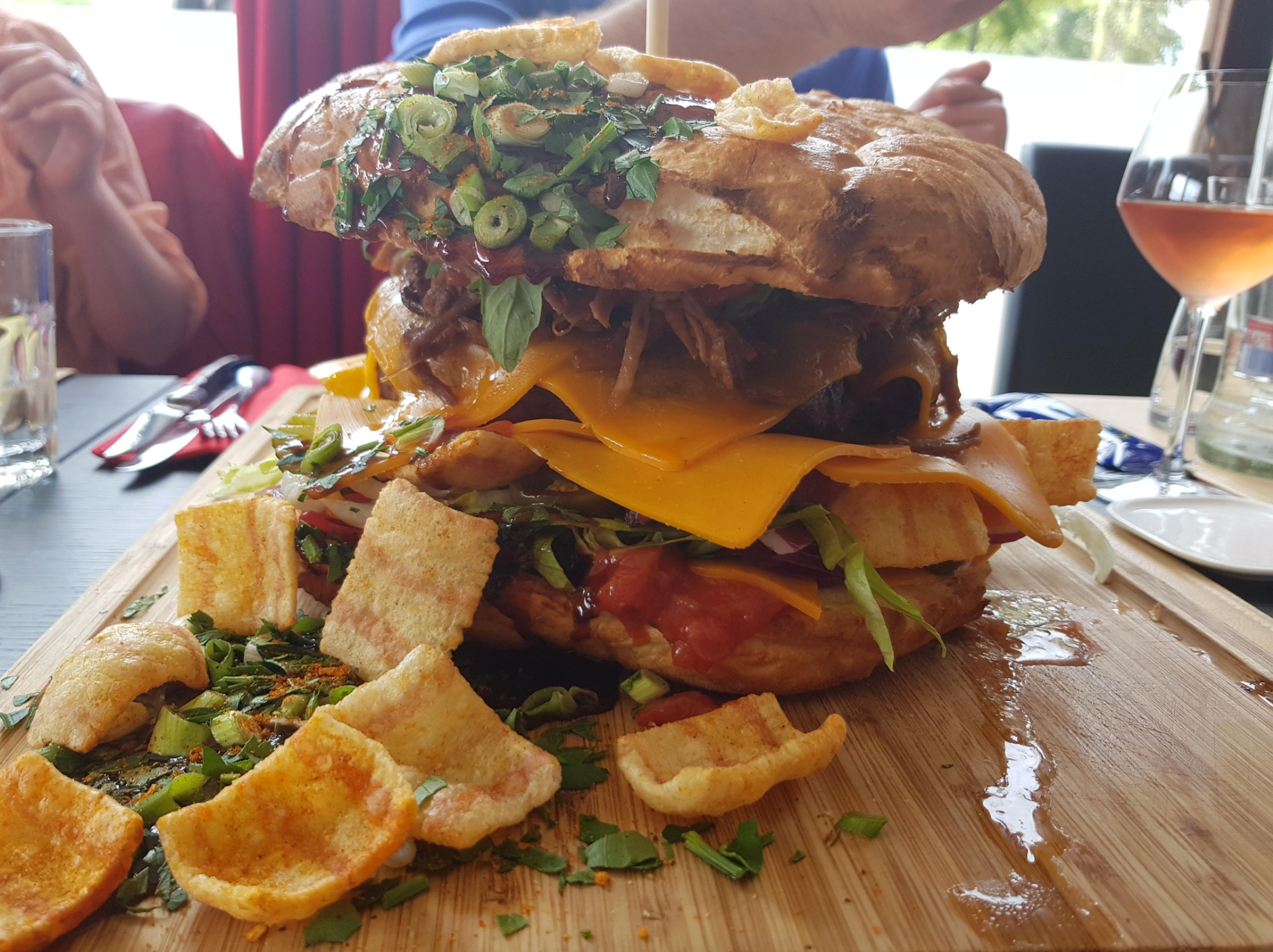 Overstuffed sandwich with cheese, greens, pulled meat, topped with crispy snacks, served on a wooden board