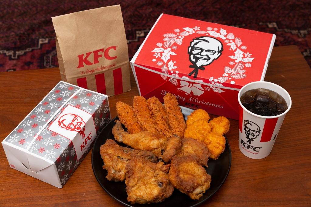 Plate with fried chicken, strips, nuggets, alongside KFC-branded boxes, a drink, and a bag on a table