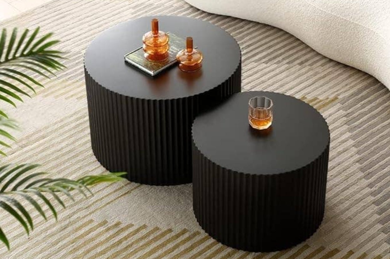 Two round, ribbed side tables with decorative items on top, including a glass and a book, set on a patterned rug near a plant