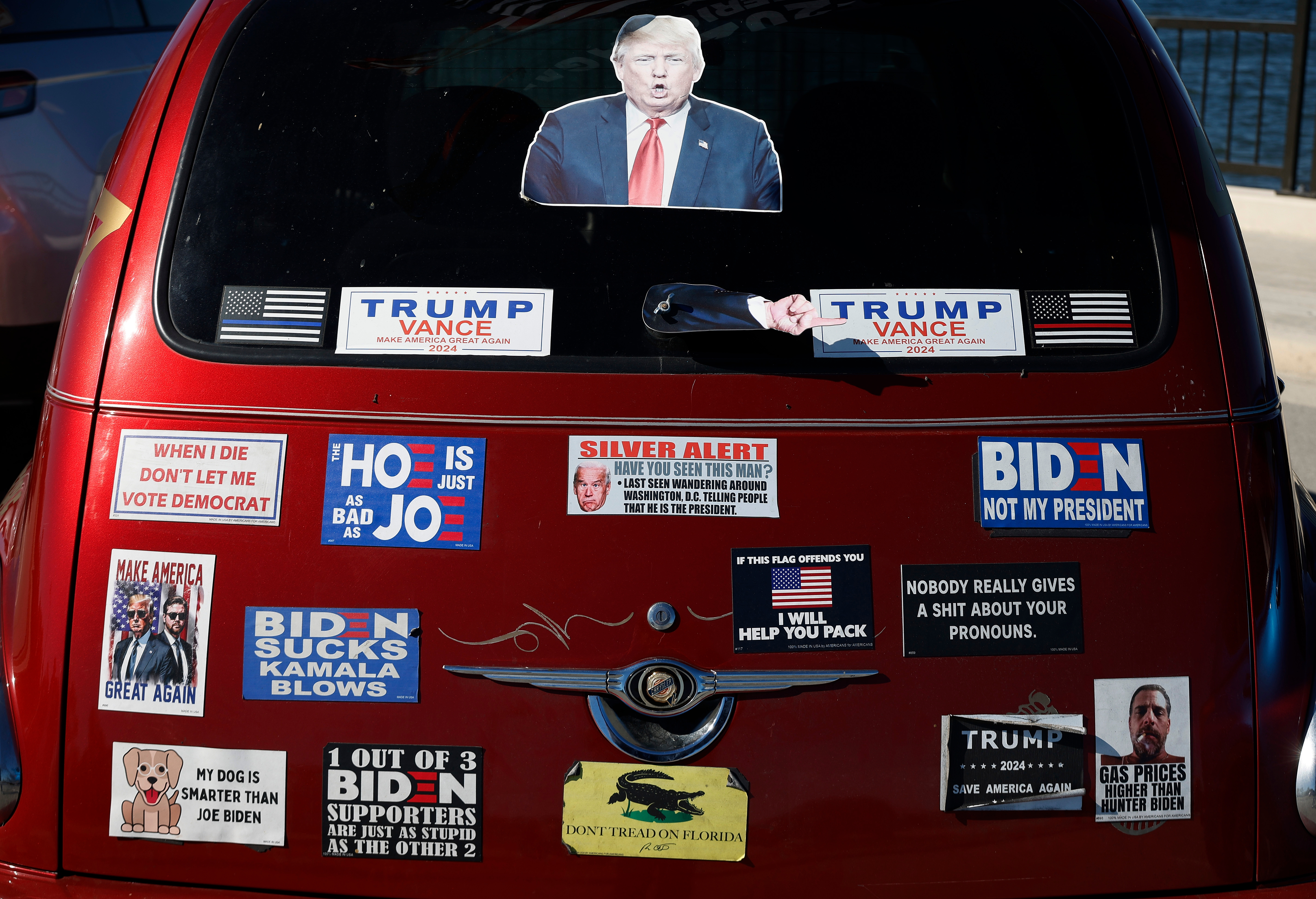 Car rear with various political bumper stickers, including satirical and critical messages about different political figures and themes
