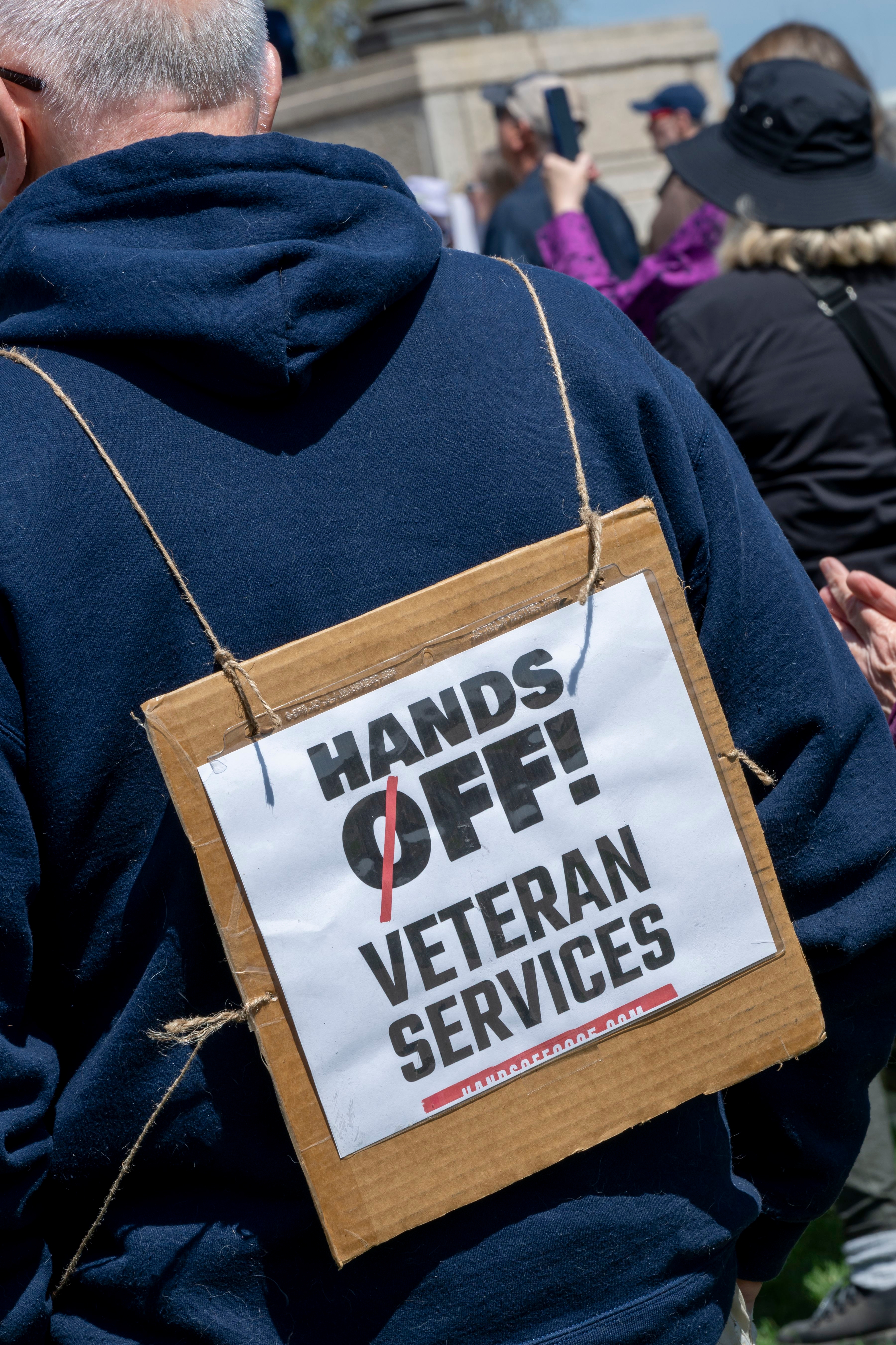 Person wearing a sign saying &quot;Hands Off! Veteran Services&quot; at a protest rally