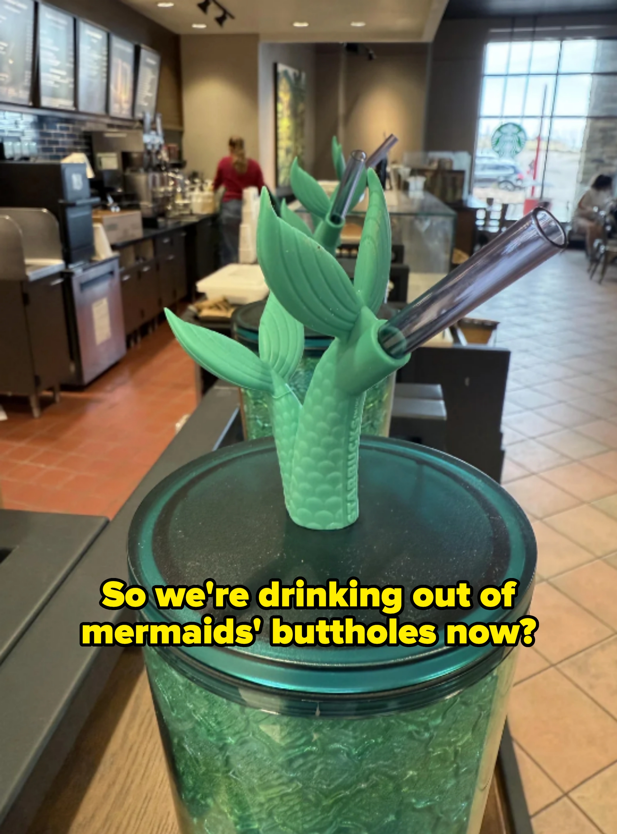 Mermaid tail-shaped straw holders atop cups at a coffee shop counter, captioned "So we're drinking out of mermaids' buttholes now?"