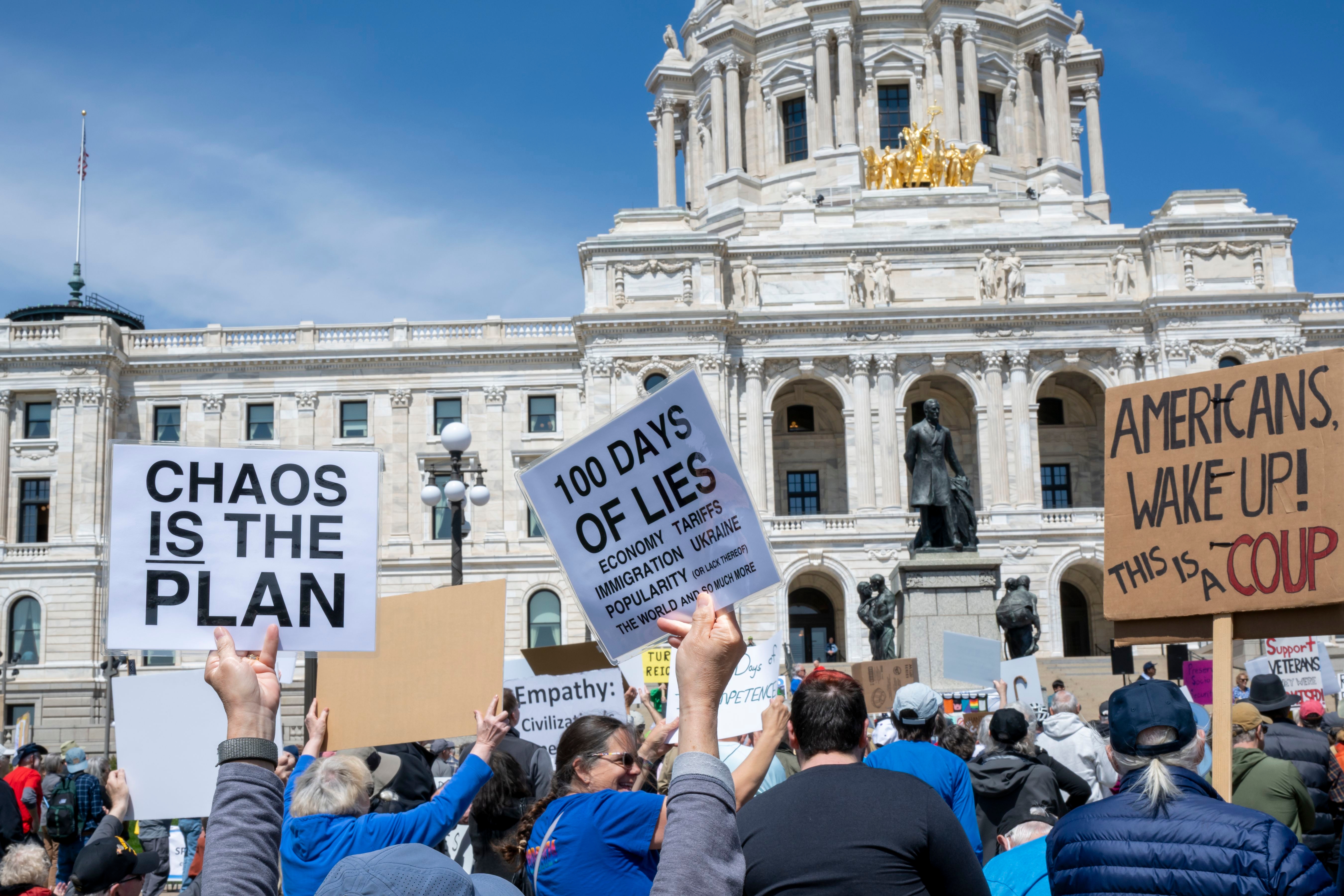 People at a protest hold signs with slogans like &quot;Chaos is the Plan&quot; and &quot;Americans, Wake Up!&quot; outside a government building