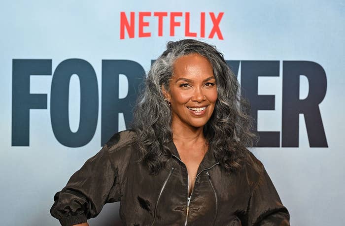Smiling woman with long, wavy hair, wearing a sleek, dark jumpsuit at a Netflix event backdrop