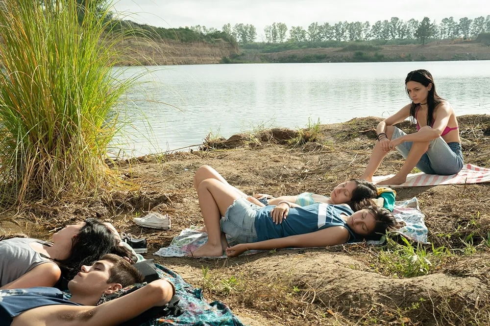 Grupo de jóvenes descansando junto a un lago, algunos sentados y otros acostados sobre mantas en la hierba