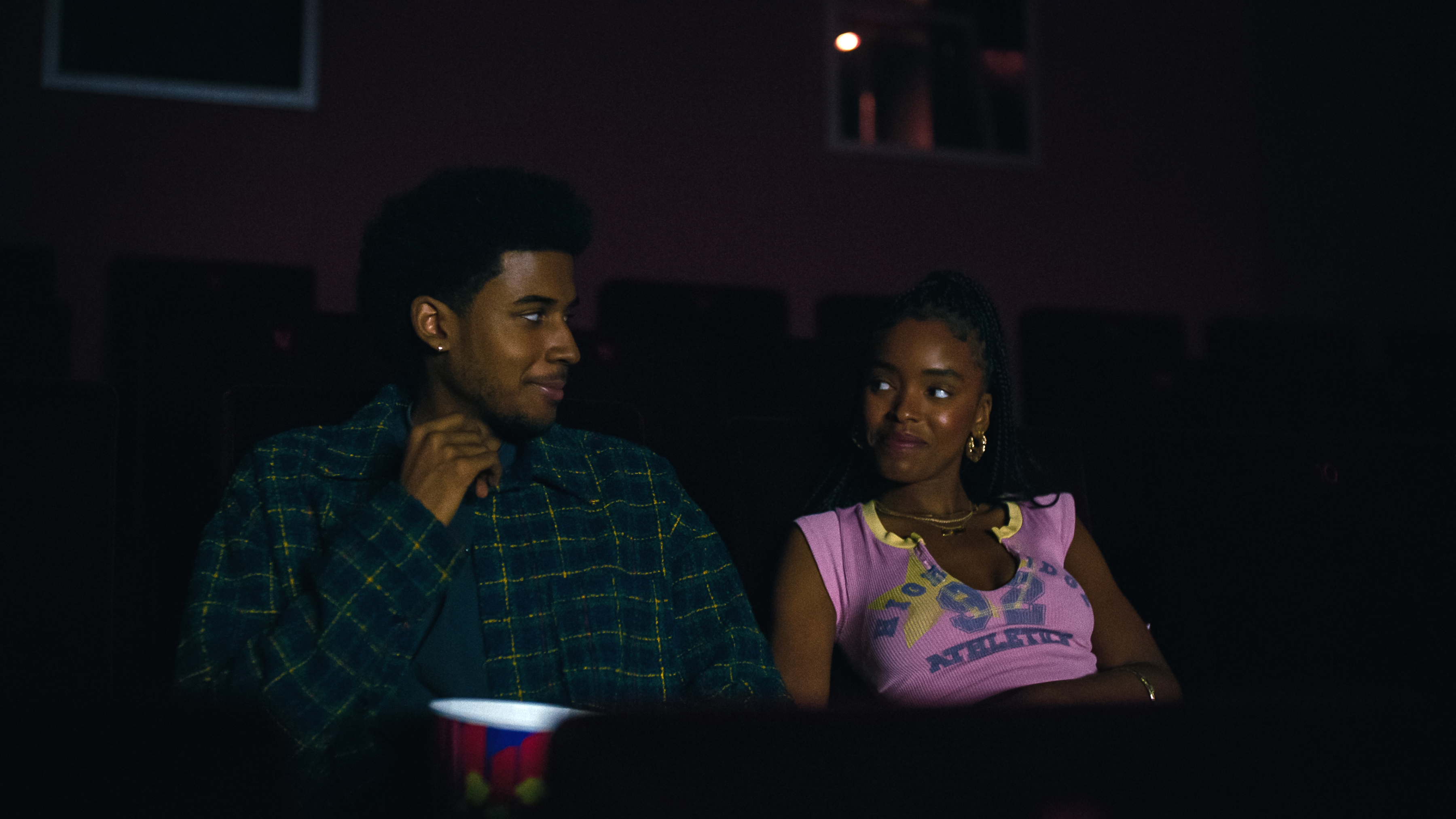 Two people sit in a dark theater, exchanging smiles. One wears a patterned shirt, the other a graphic tee. A popcorn tub is visible