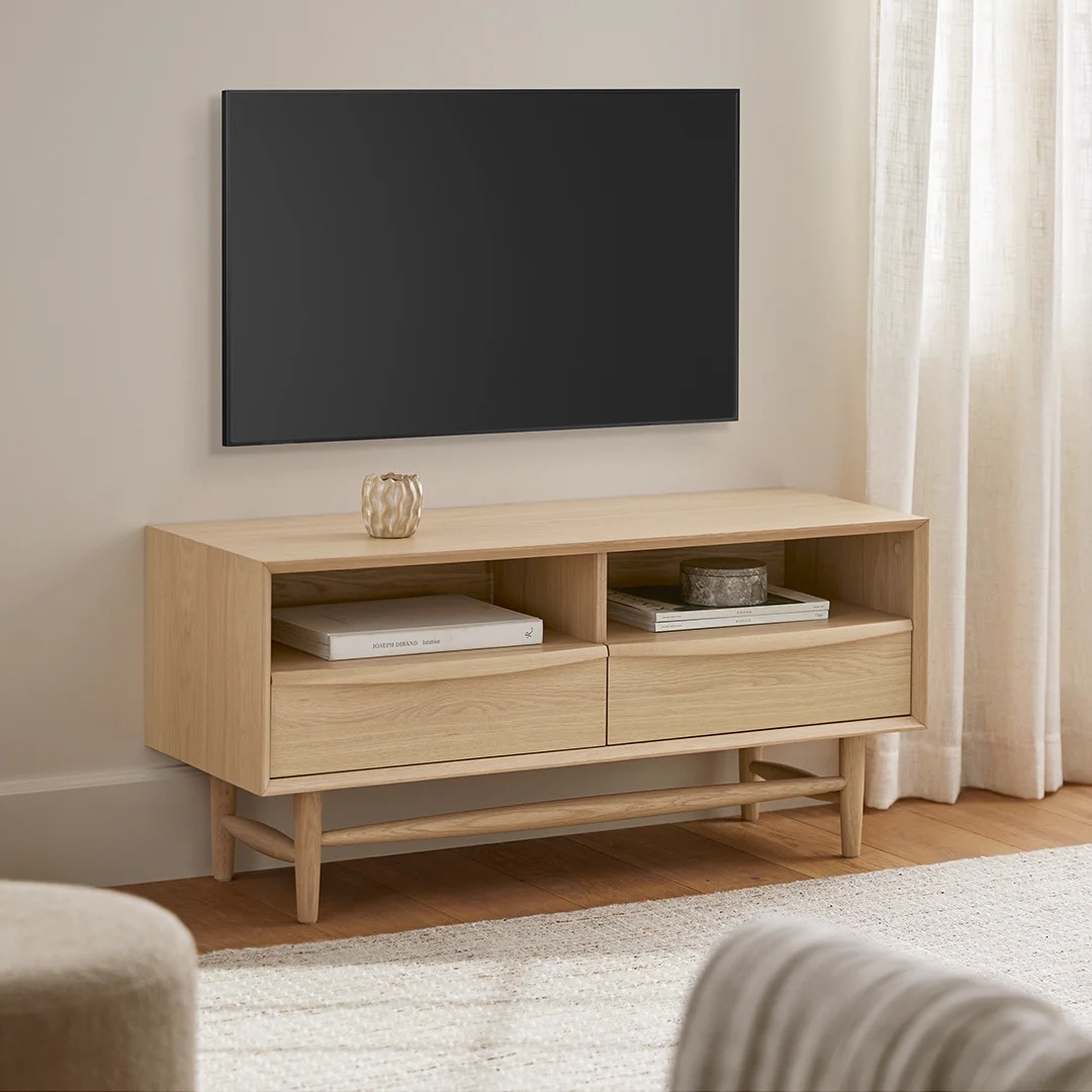 Modern wooden TV stand with shelves and drawers, displaying a decor item and book. Wall-mounted TV above. Neutral living room setting