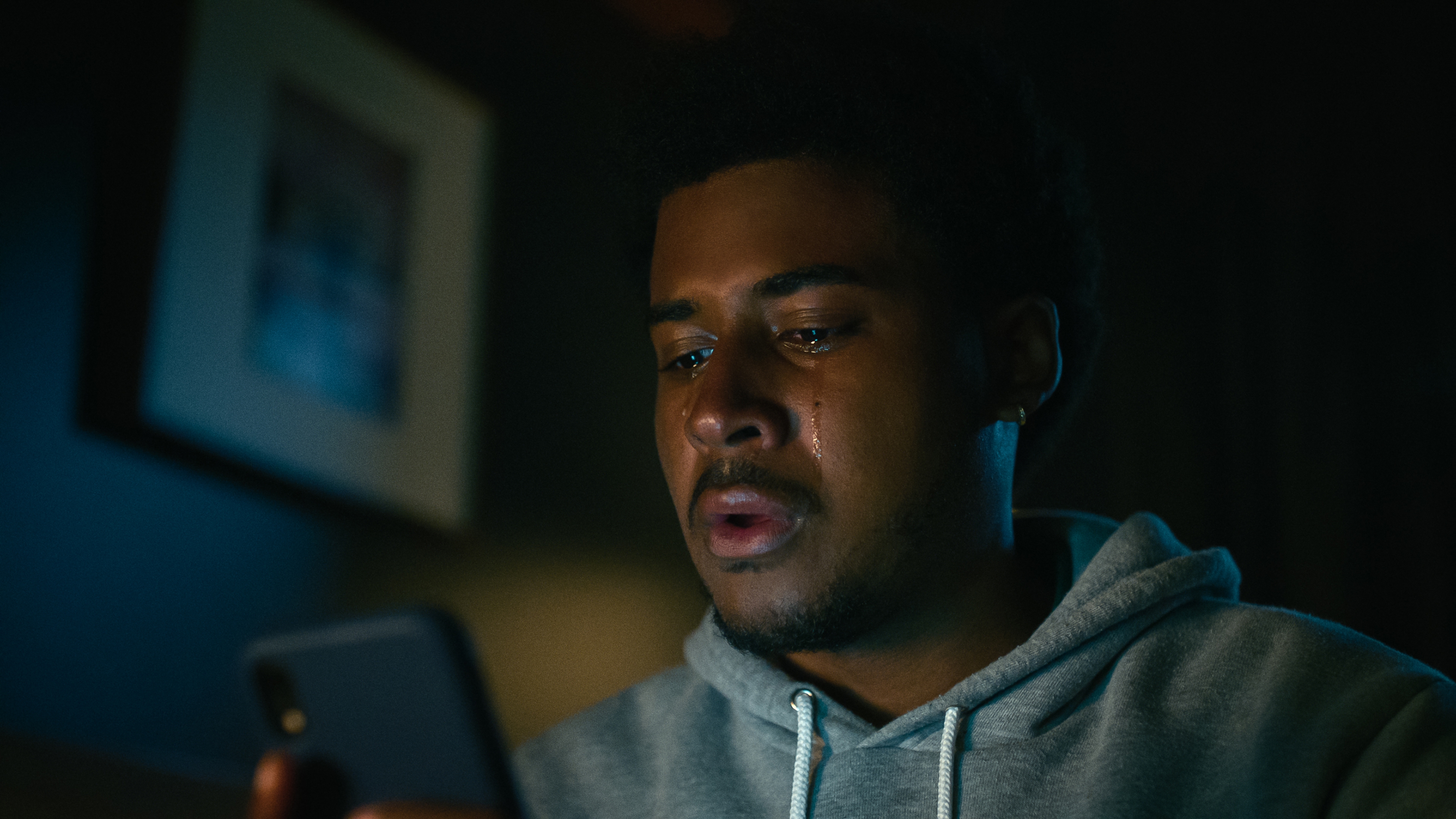 Person in a hoodie looks intently at their phone, tears on their face, suggesting emotional news or a heartfelt moment