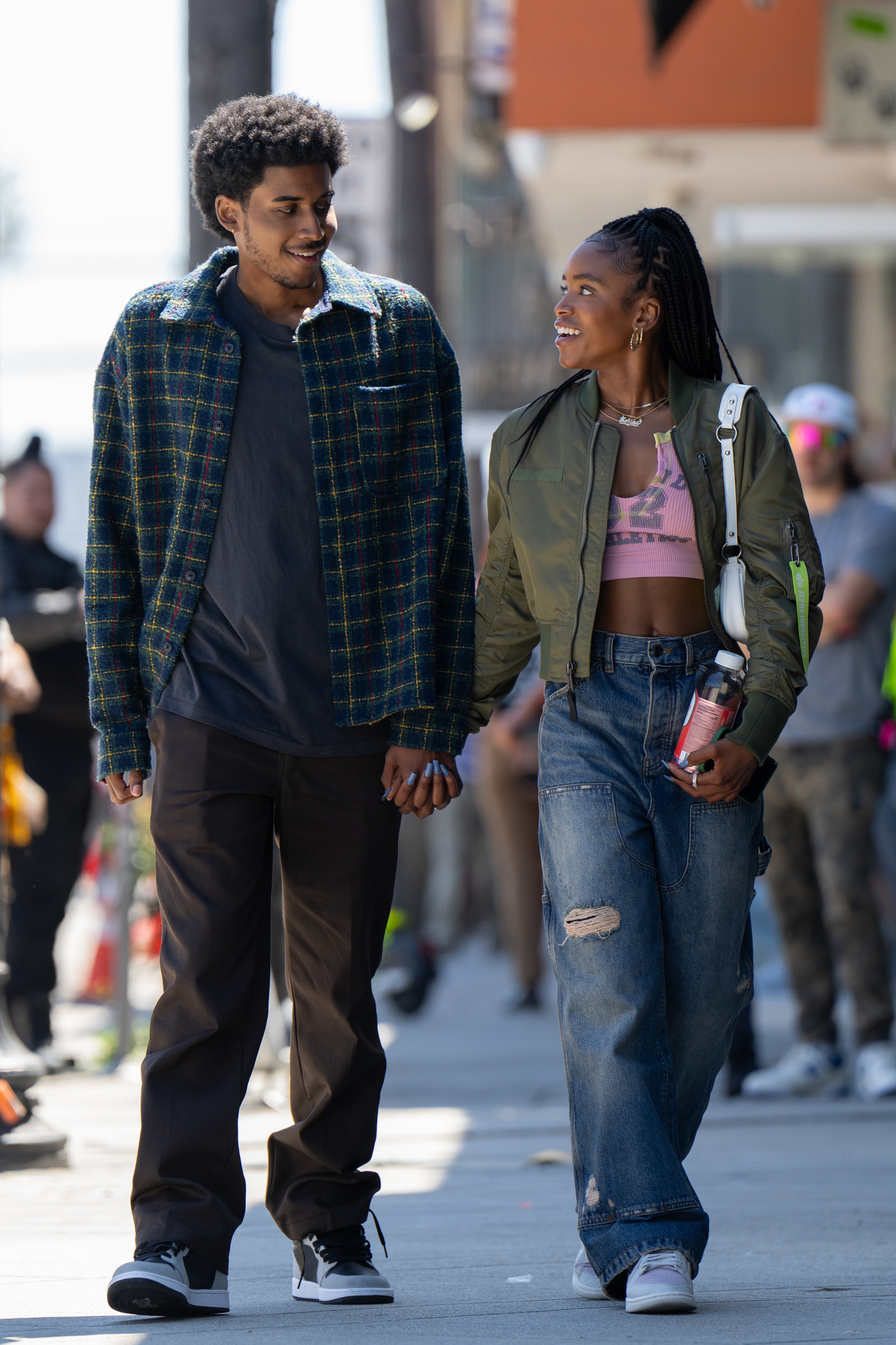 Two people walking hand in hand on a city street, smiling at each other. One wears a plaid jacket, and the other wears a green bomber jacket and ripped jeans
