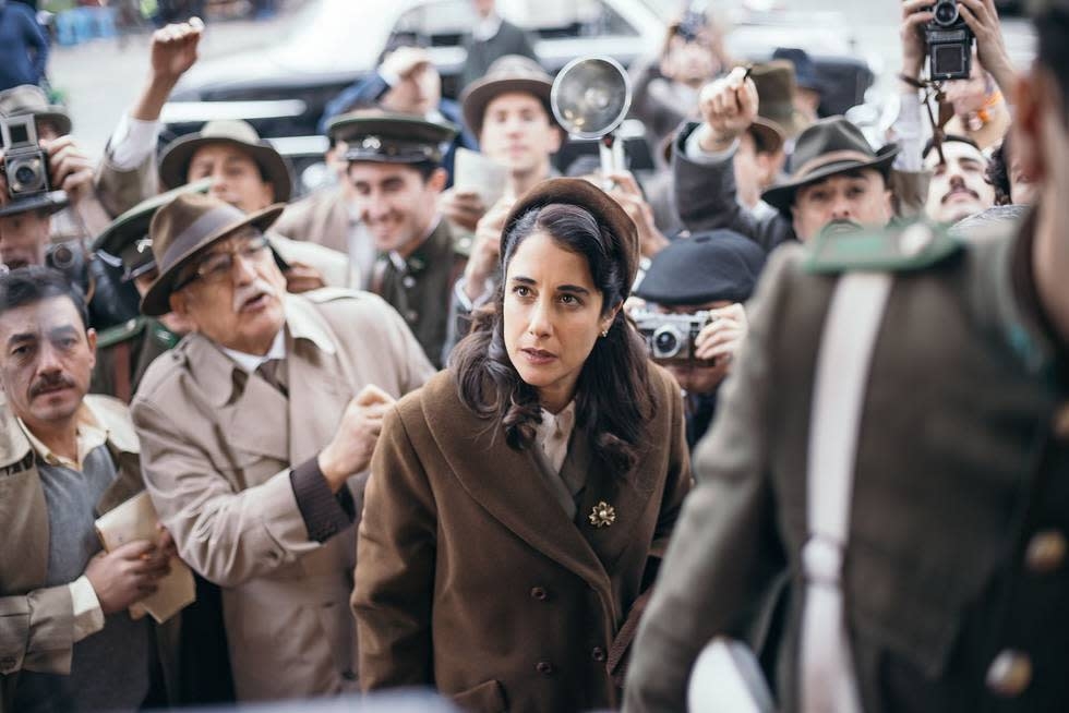 Persona rodeada de reporteros y fotógrafos, luciendo un abrigo de estilo vintage. Ambiente de prensa intensa