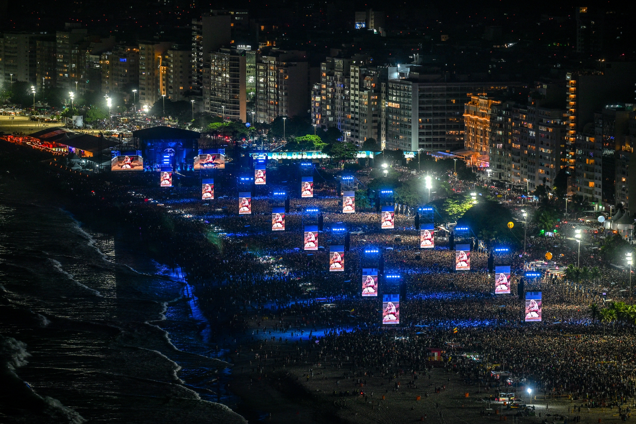 Aerial view of a large outdoor concert with illuminated screens and a packed crowd near a cityscape at night