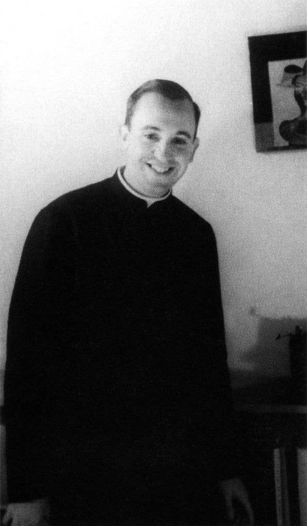 A person in priest attire smiles while standing indoors next to a wall-mounted artwork