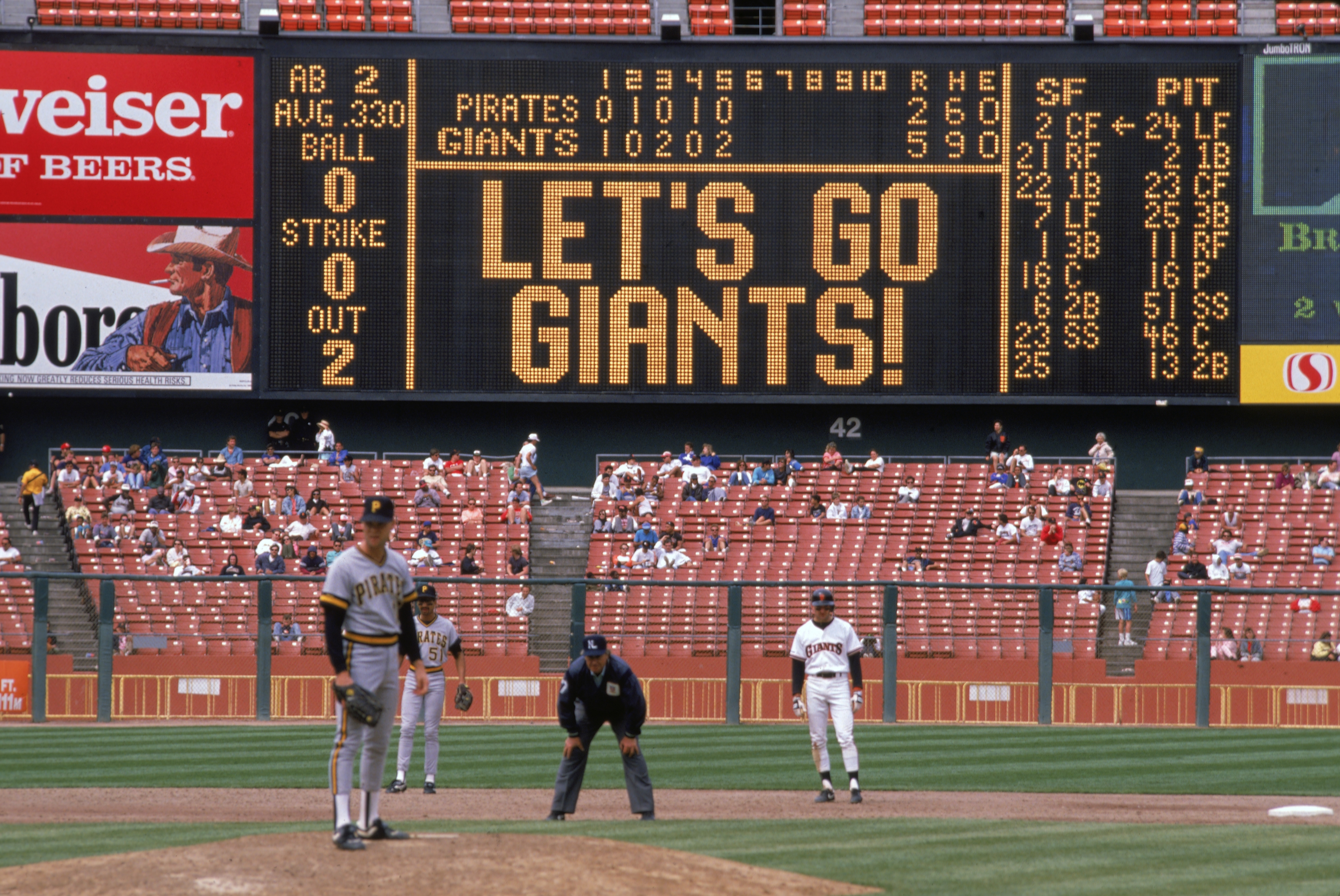 Baseball game scoreboard displays "LET'S GO GIANTS!" with teams' scores and player stats. Players and umpire on field, a few fans in stands