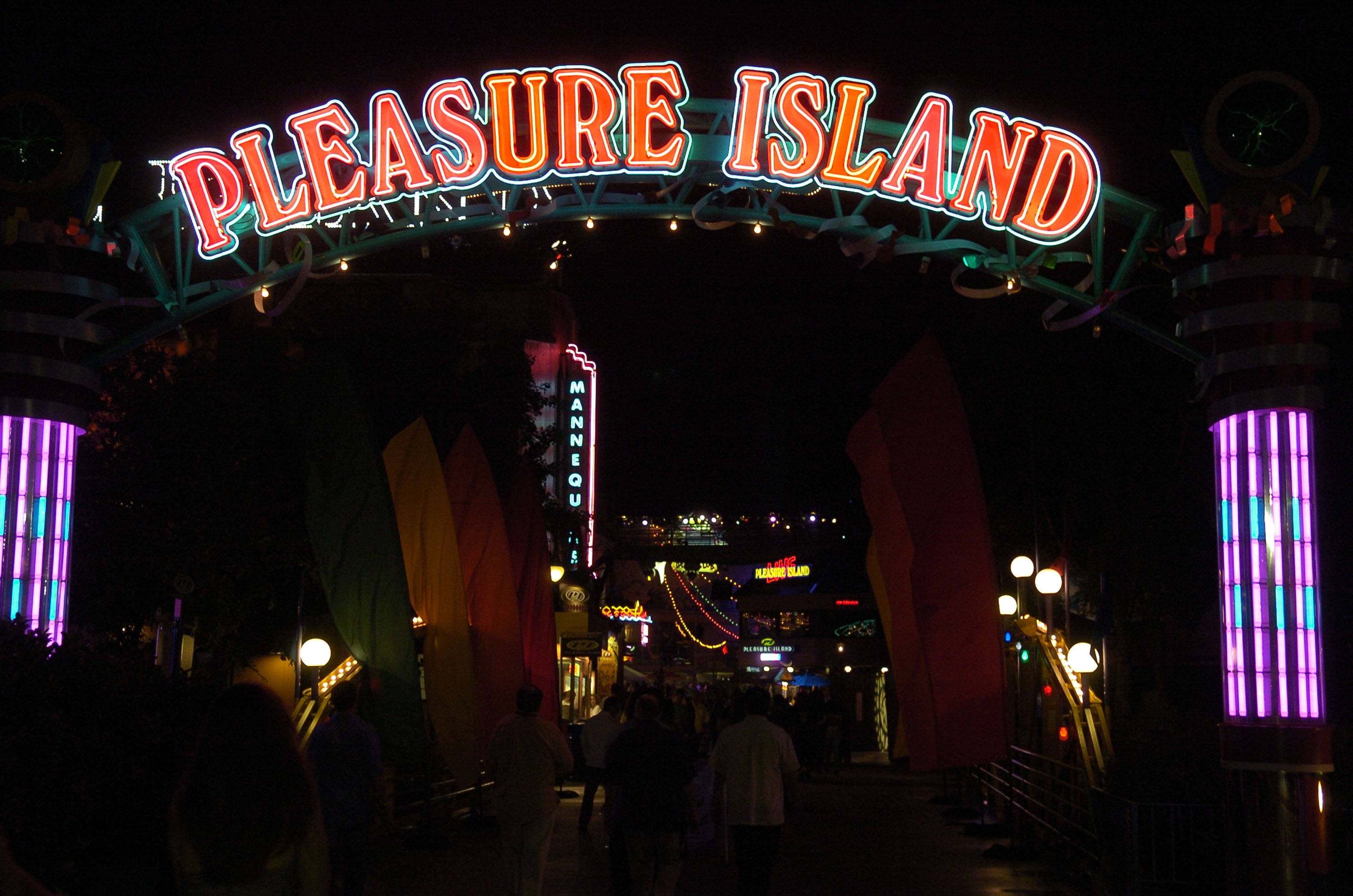 Entrance to Pleasure Island at night, illuminated by neon signs, with people walking underneath