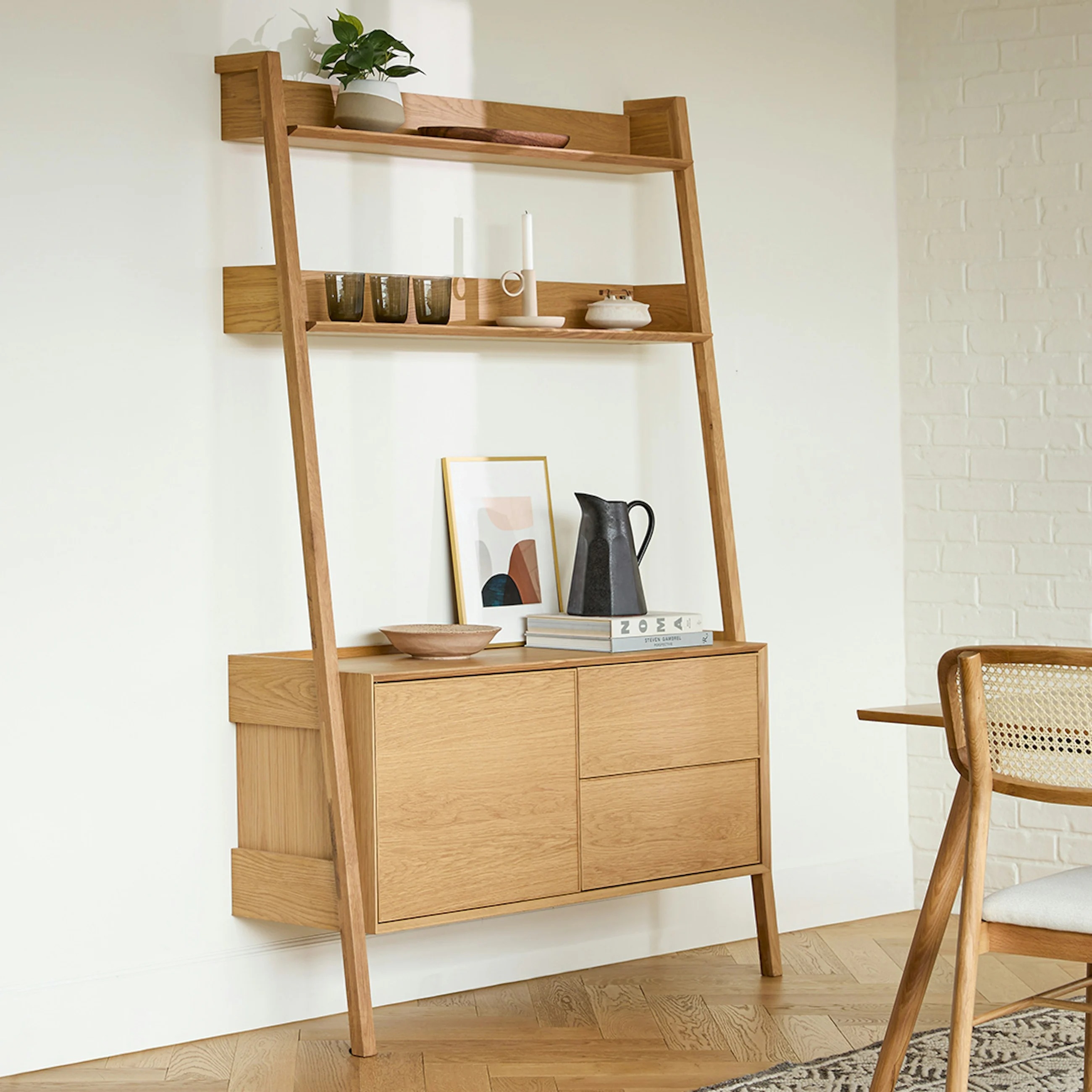 Wooden leaning ladder shelf with decorative items like a pitcher, candles, and framed art on steps; set in a modern, minimalist room