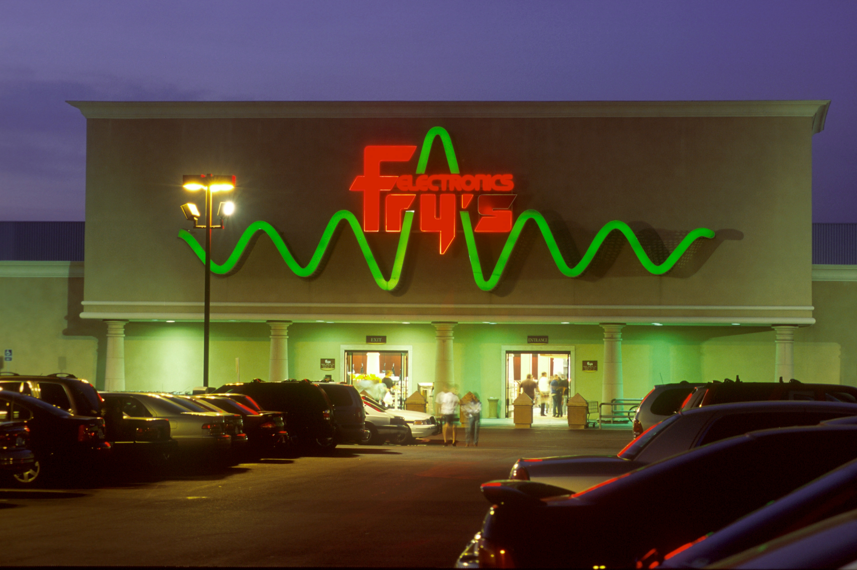 Exterior of Fry's Electronics store entrance, lit signage with distinctive green wave. Cars parked outside during evening