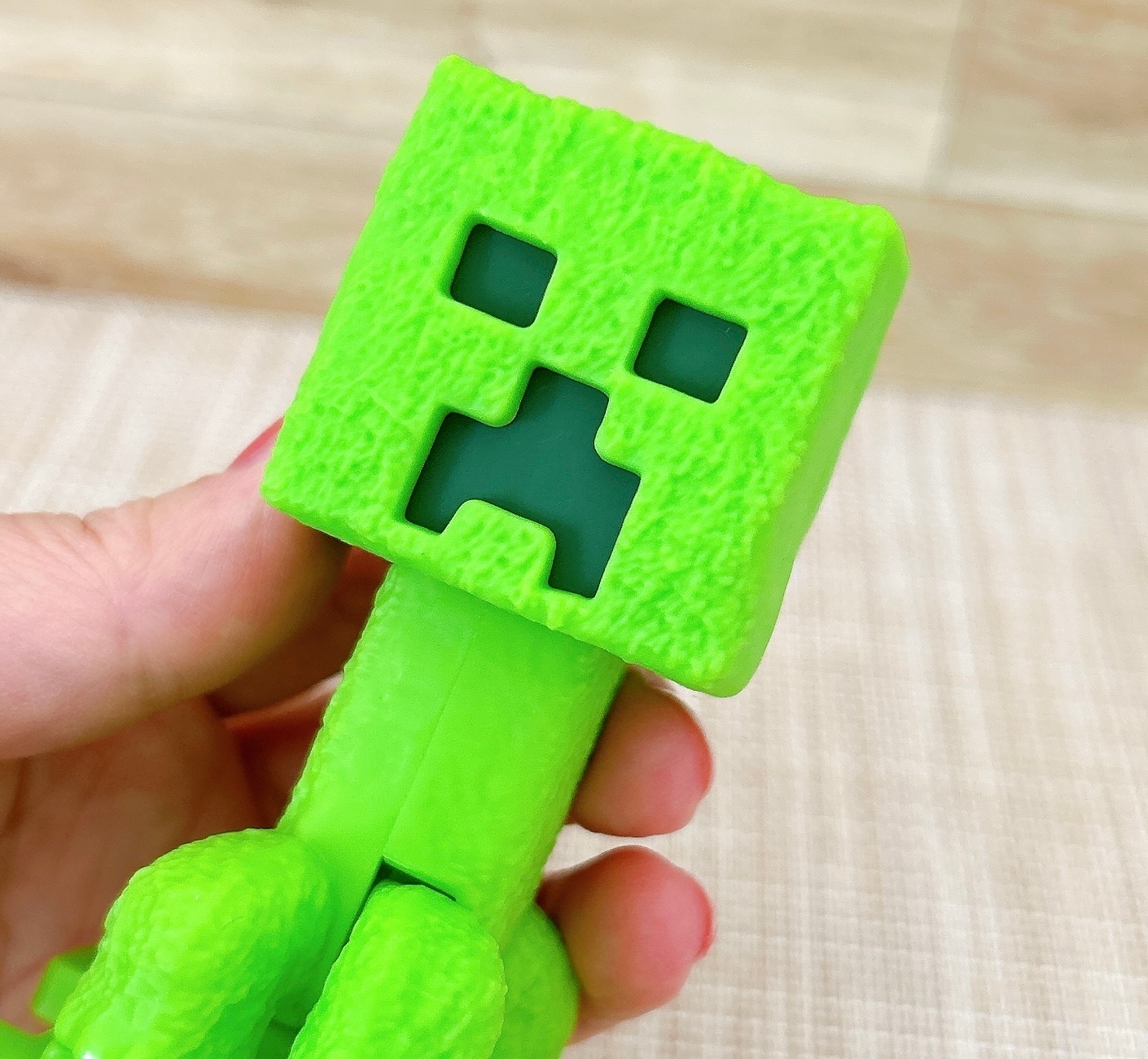 緑色のブロック状キャラクターを手に持っています。キャラクターは立方体の頭部と簡単な顔のデザインがあります。