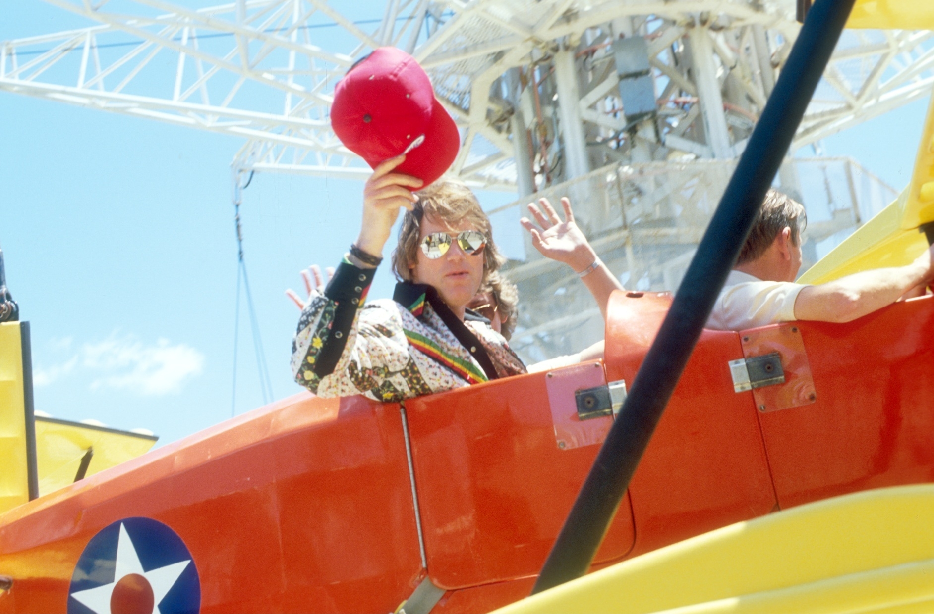 Person in aviator sunglasses and patterned jacket waves red cap from biplane with military insignia. Background features large structure