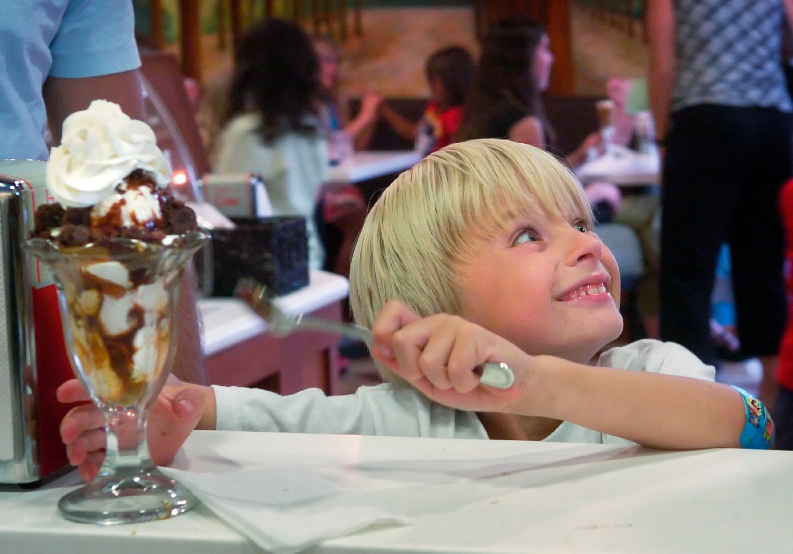Child smiles eagerly at an ice cream sundae topped with whipped cream, holding a spoon, in a busy ice cream parlor