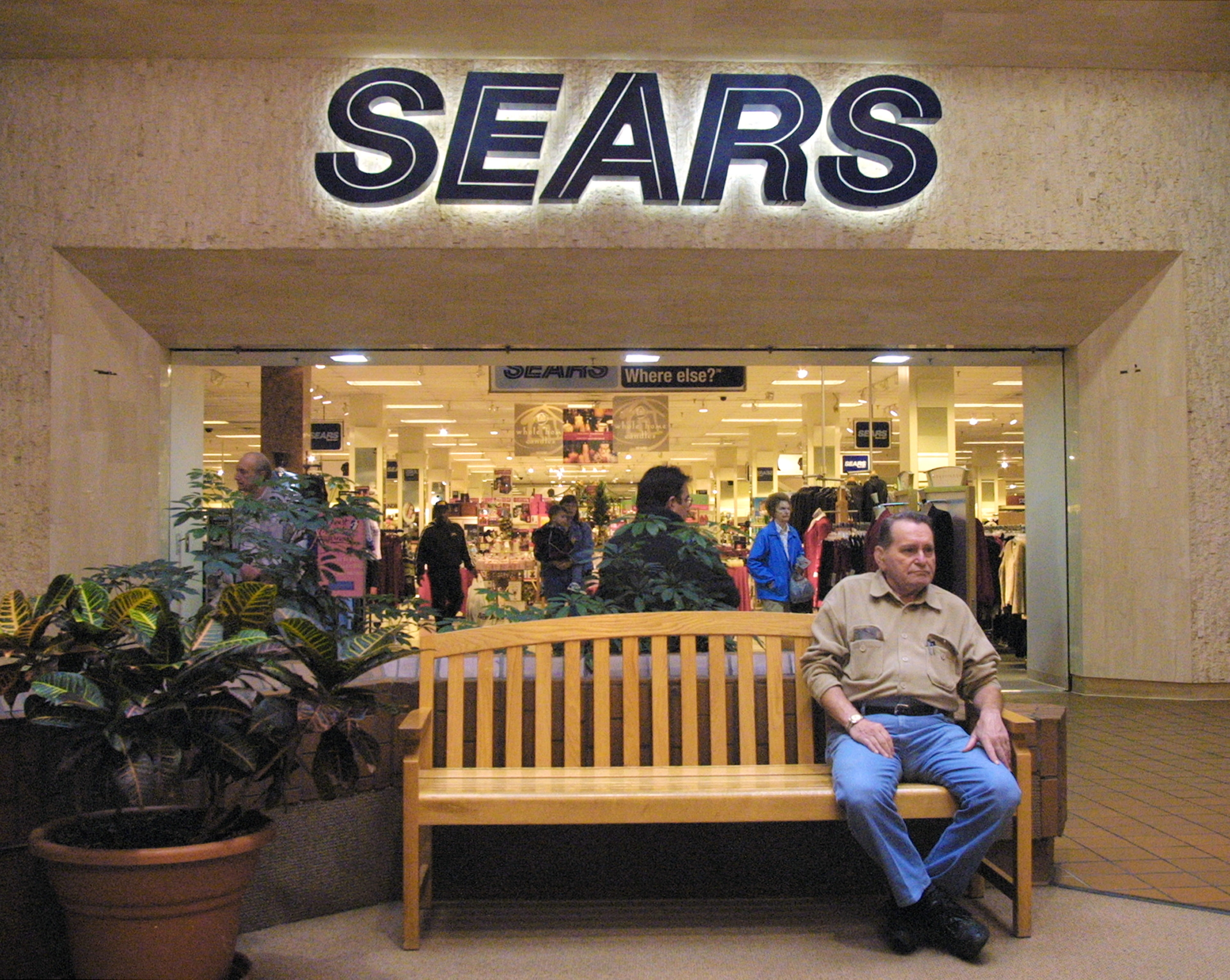 A man sits on a bench outside a busy Sears store, located within a shopping mall