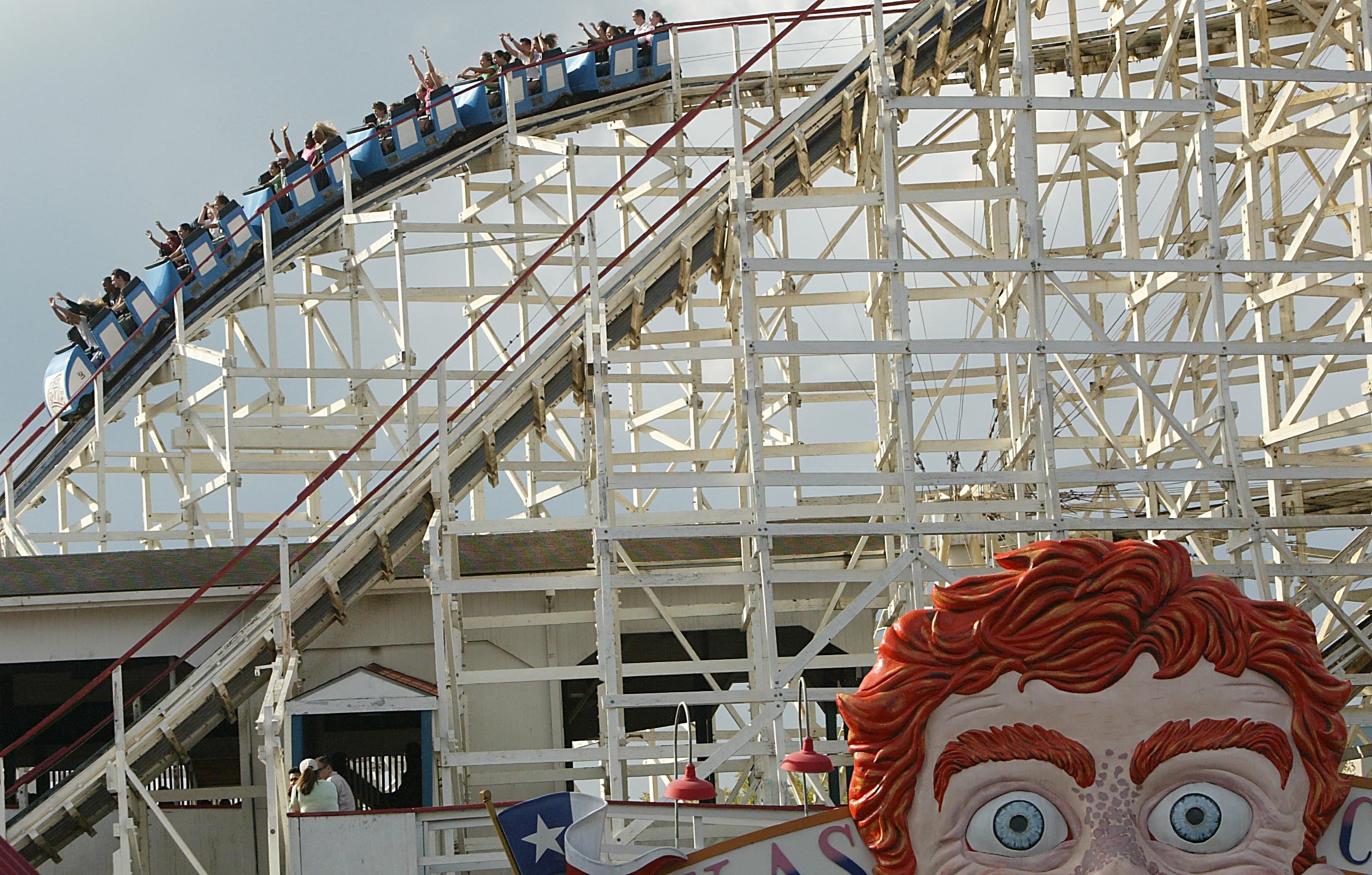 People ride a large wooden roller coaster, raising their hands in excitement. A giant face sculpture is partially visible in the foreground