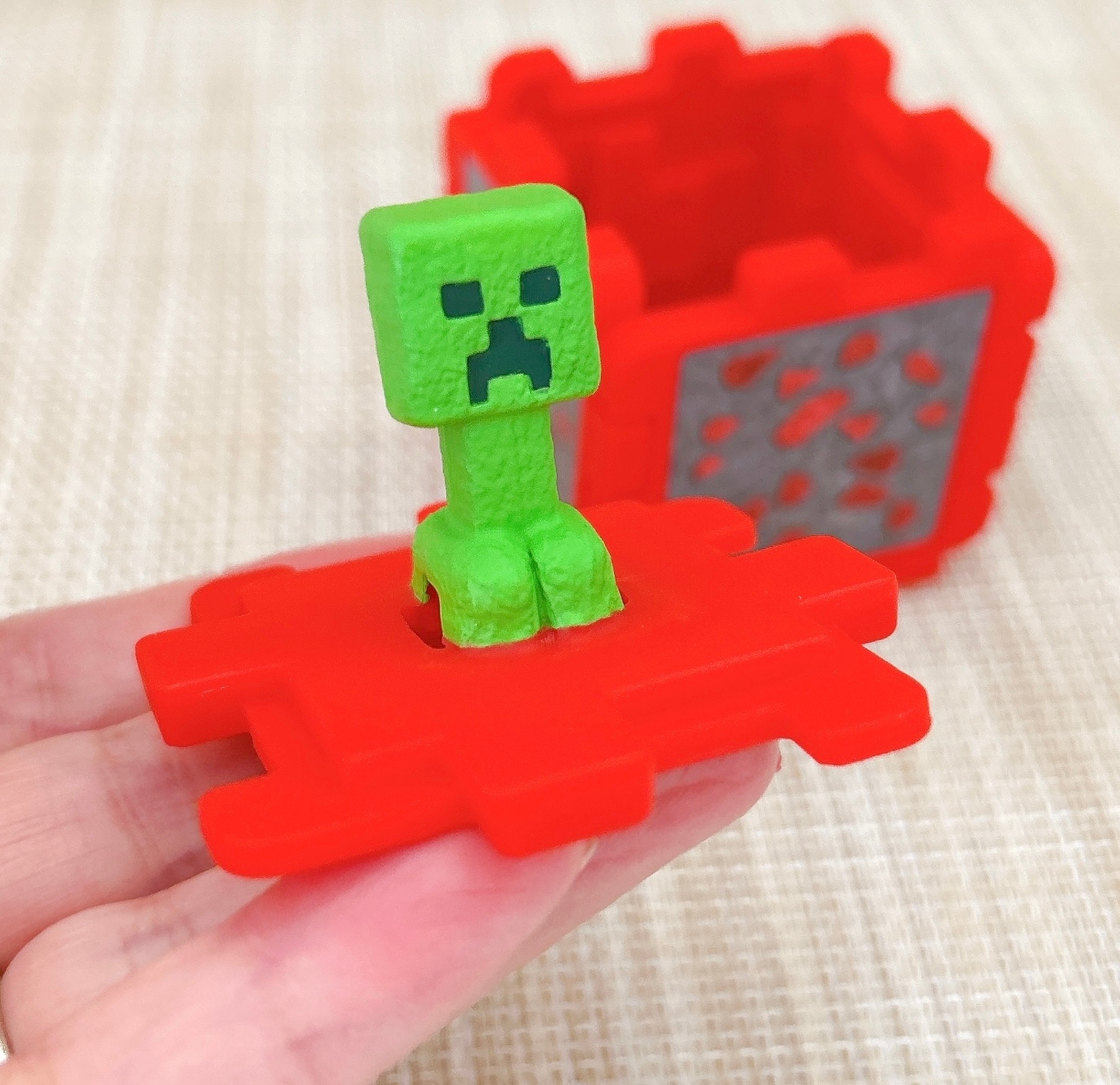 手に持たれた立体パズルの一部には、緑色のブロックキャラクターが乗っています。後ろには赤い箱の一部が見えます。