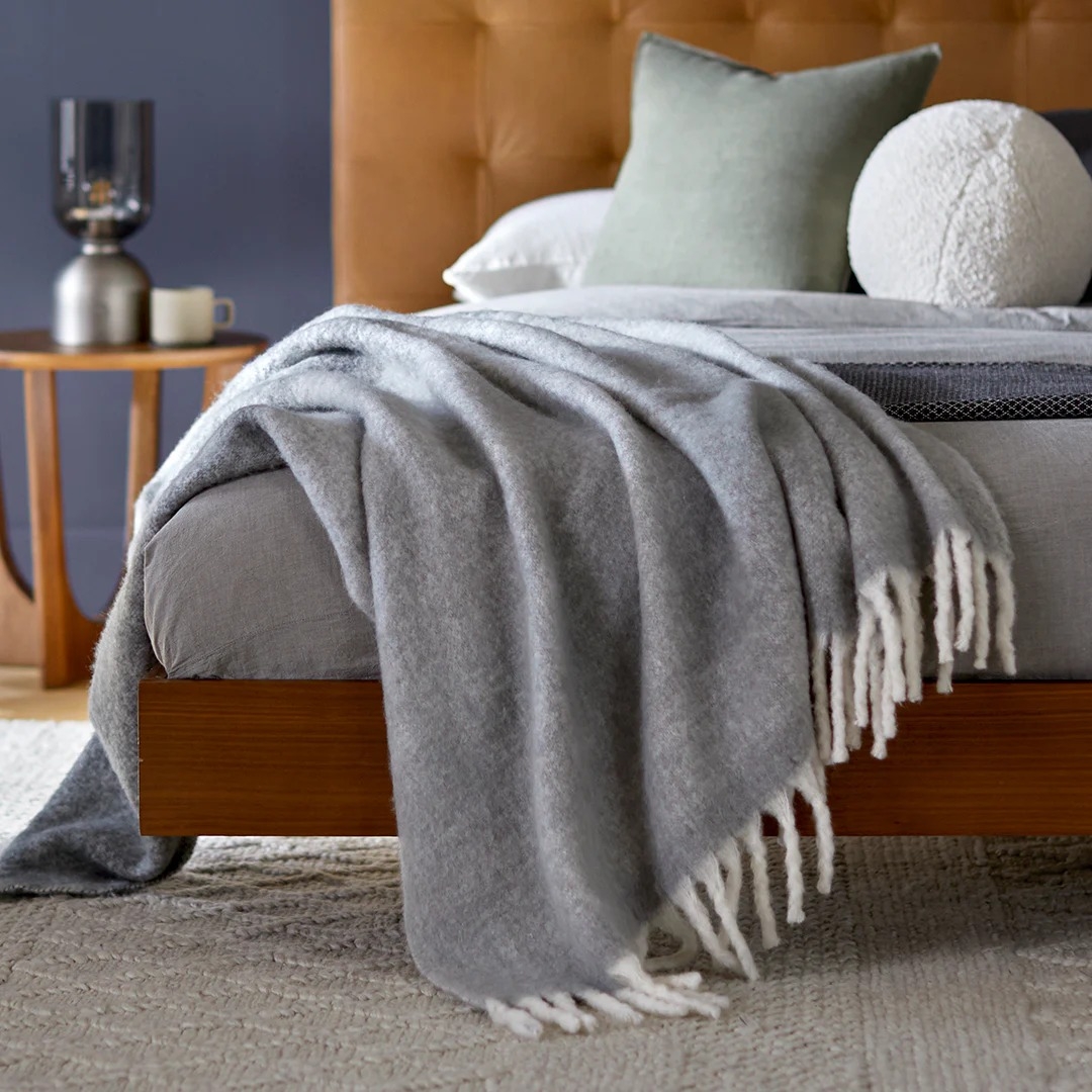 Modern bedroom with a wooden bed frame, gray throw blanket, and neutral pillows. A side table holds a lamp and a cup, creating a cozy atmosphere