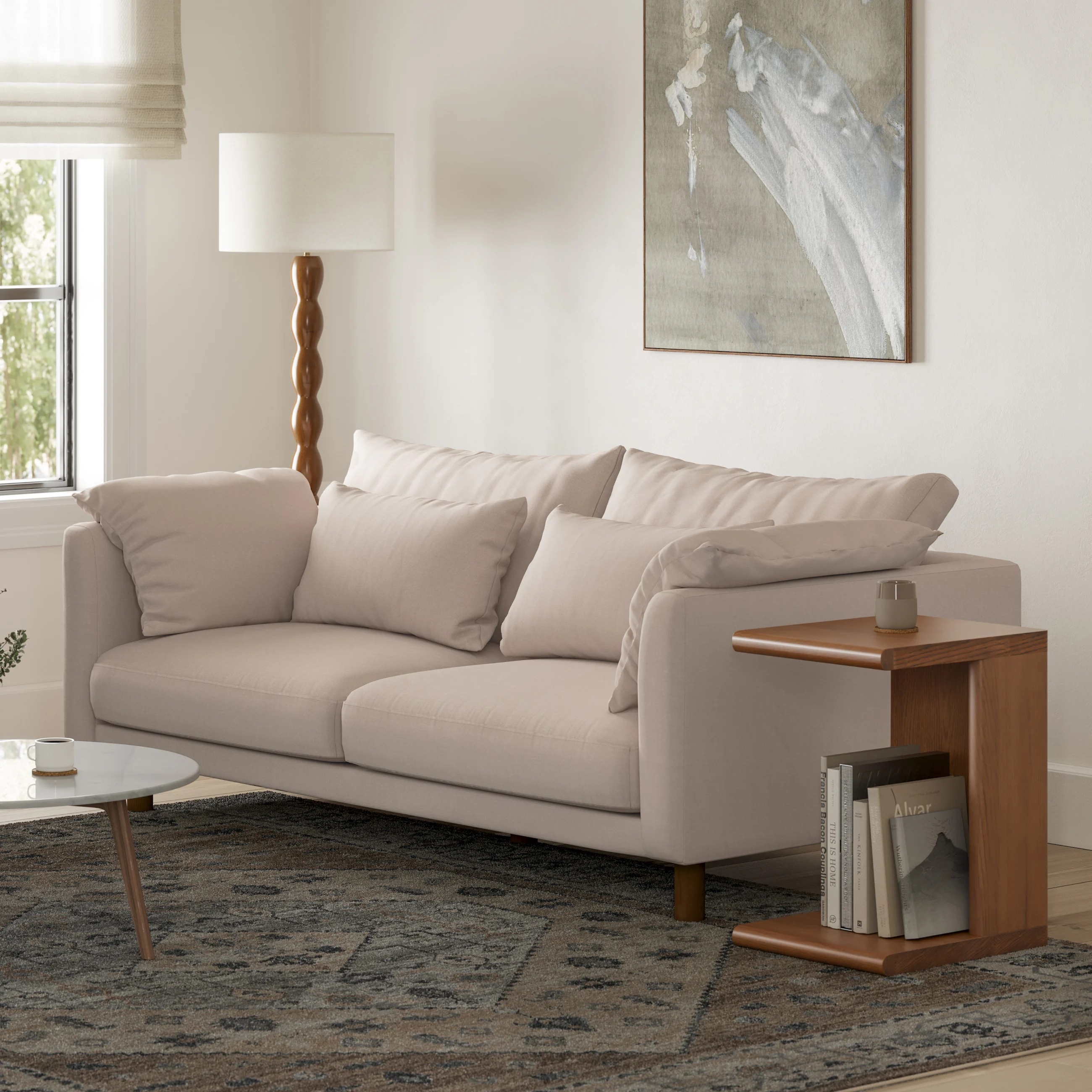 Modern living room with a plush sofa, round coffee table, and a small wooden side table holding books. Lamp and abstract art in the background