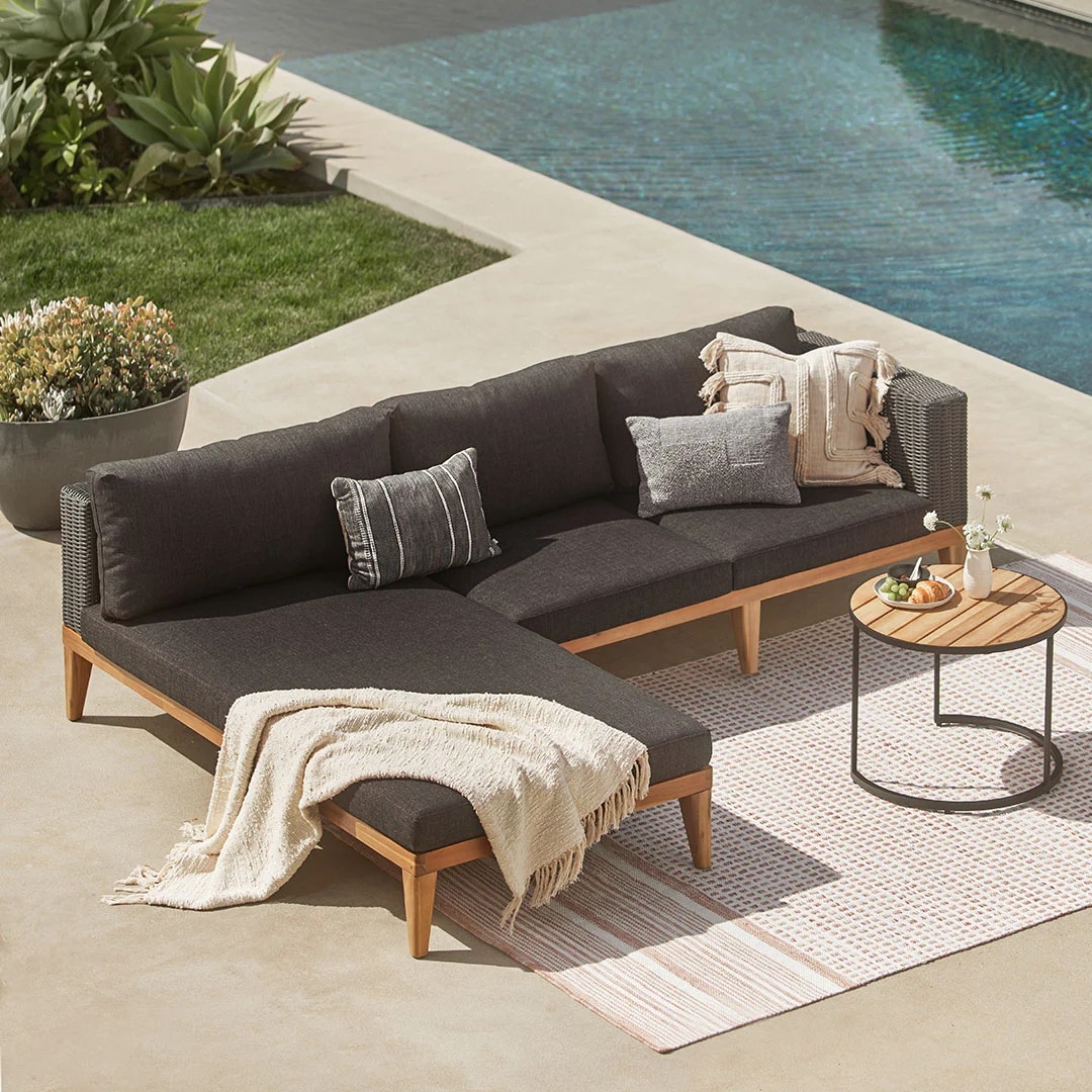 Outdoor patio with dark cushioned sectional sofa, throw pillows, and a blanket, placed on a textured rug beside a round table and poolside setting