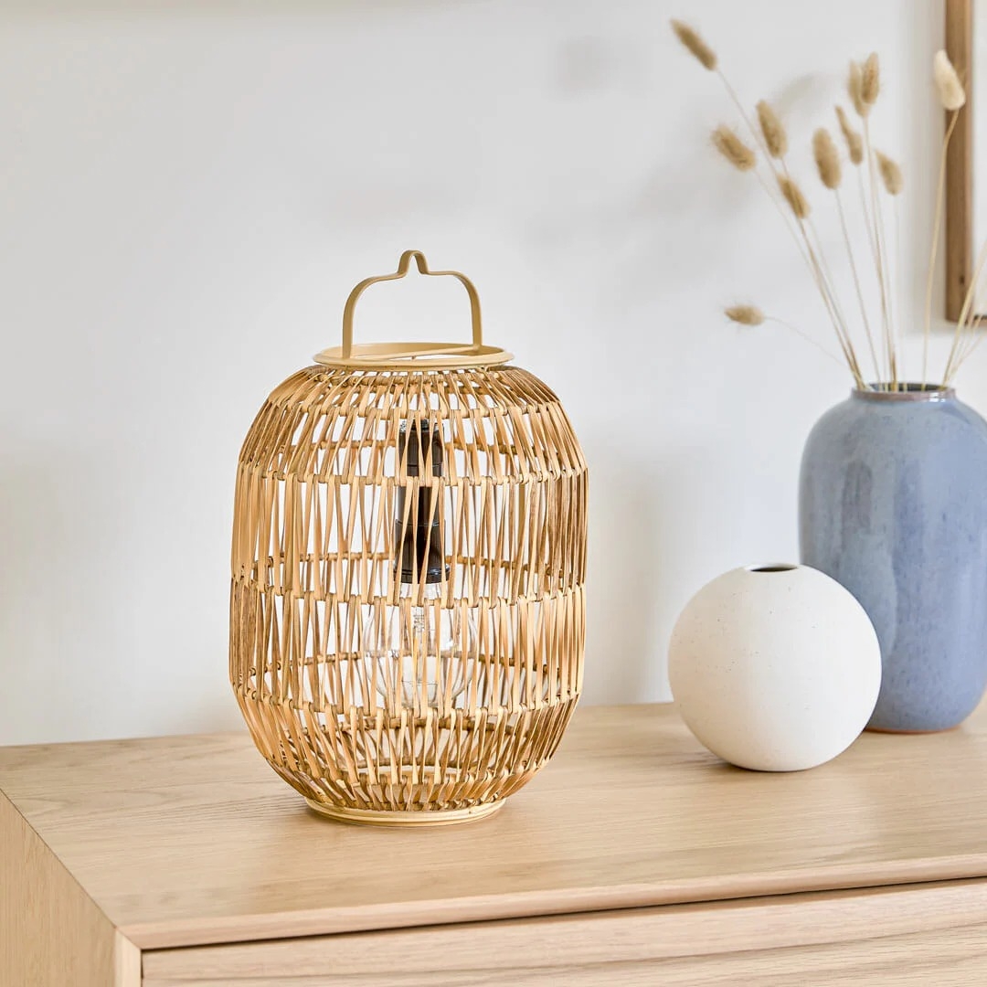 A decorative wicker lantern sits on a wooden dresser next to a white round vase and a blue vase with dried grass